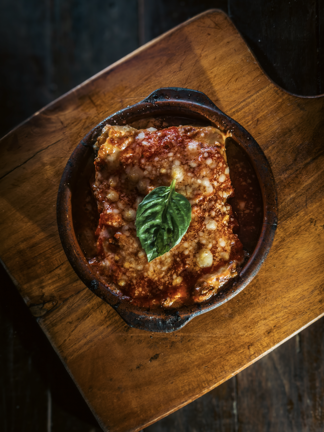 Baked pasta with cheese and tomato sauce garnished with basil leaf, served in a rustic clay dish on wooden table.