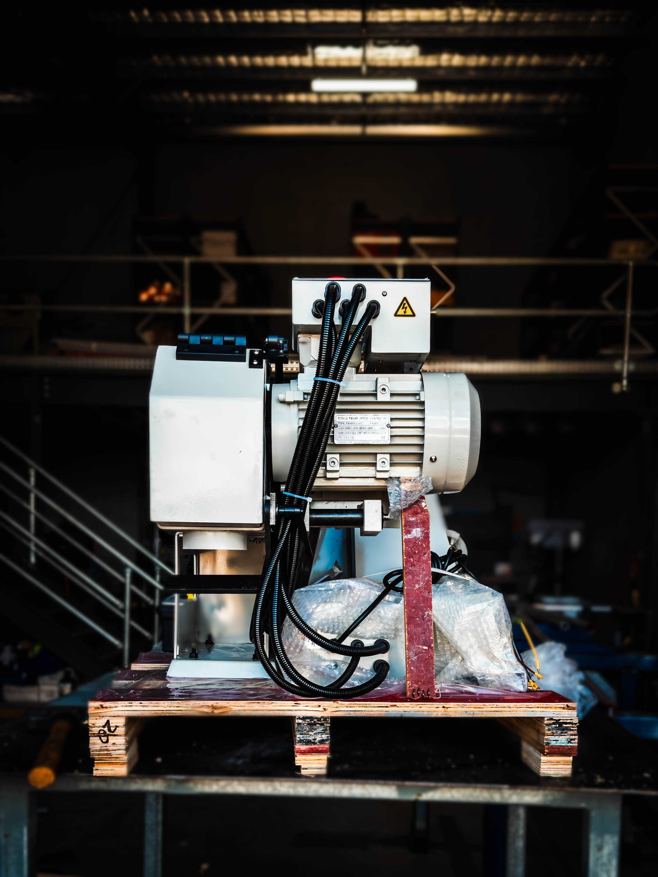 Industrial machinery or motor with wires mounted on a wooden pallet in a warehouse or factory setting.