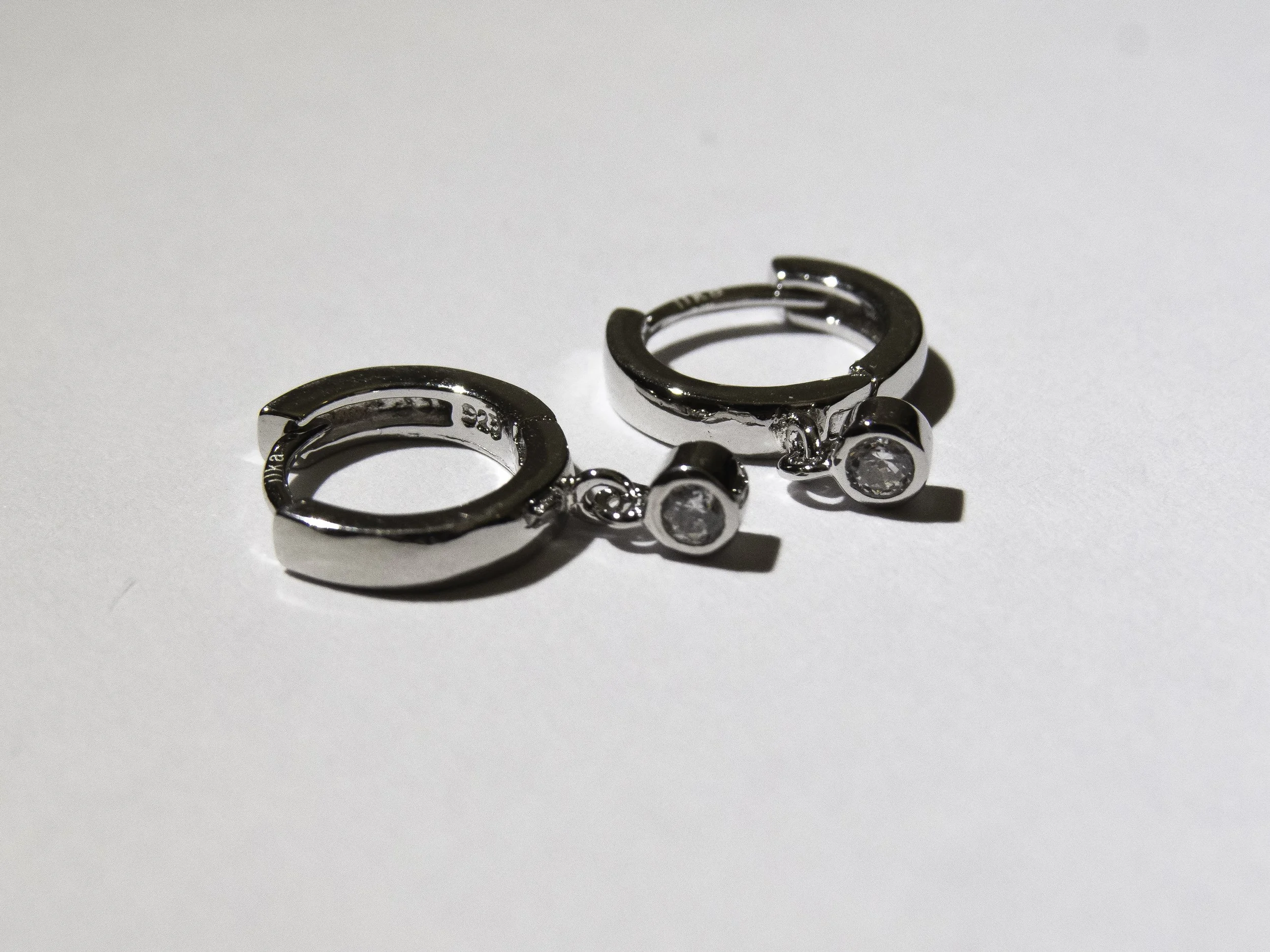 A pair of silver earrings with clear round gemstones placed on a white surface.