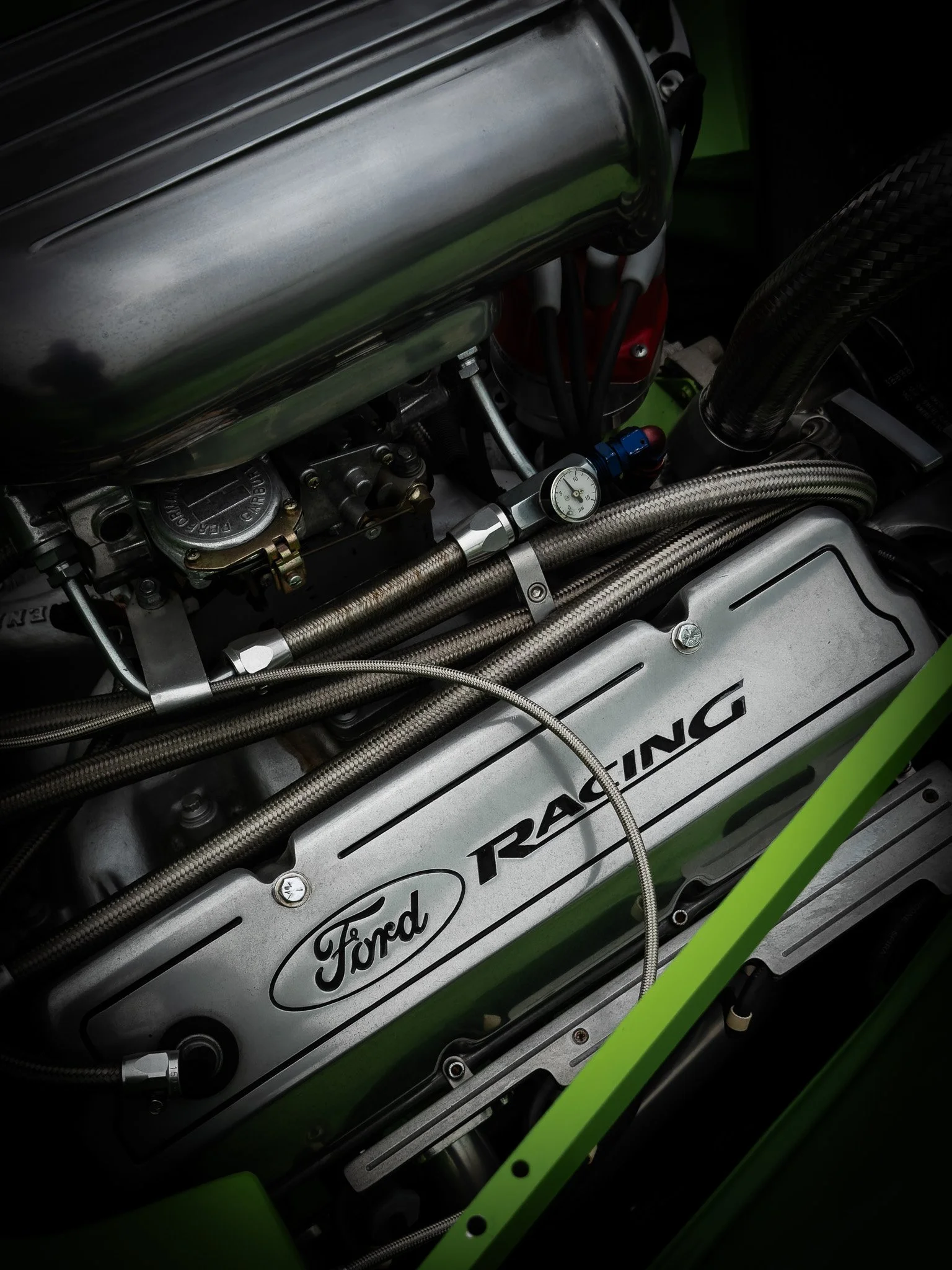 Close-up of a racing car engine with visible Ford and Roush branding, metallic components, braided hoses, and a pressure gauge.
