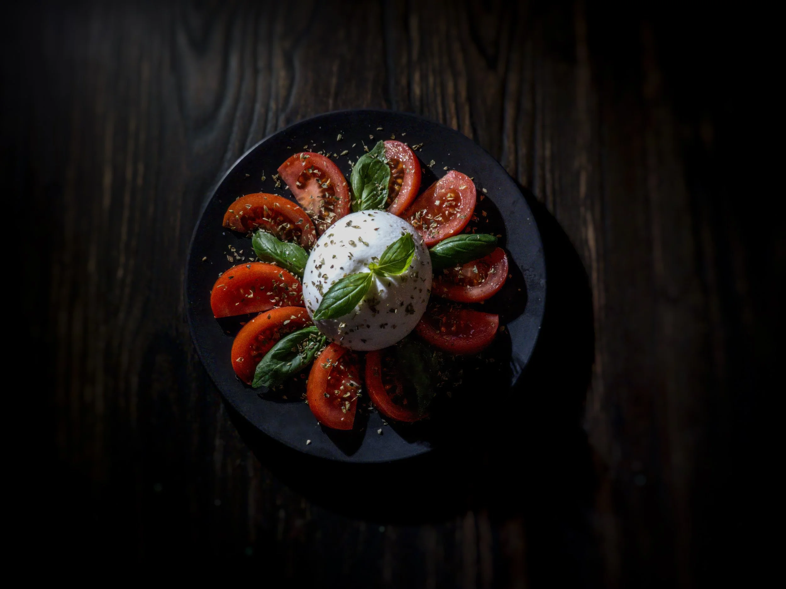 Caprese salad with sliced tomatoes, fresh basil leaves, mozzarella ball, sprinkled with herbs on a dark plate.