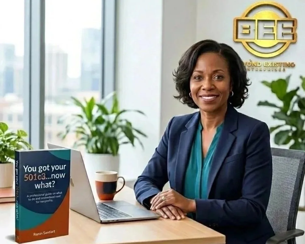 A woman sitting at a desk in an office, smiling at the camera, with a book titled "You got your 501c3...now what?" on the desk, a laptop, a cup, and green plants in the background.