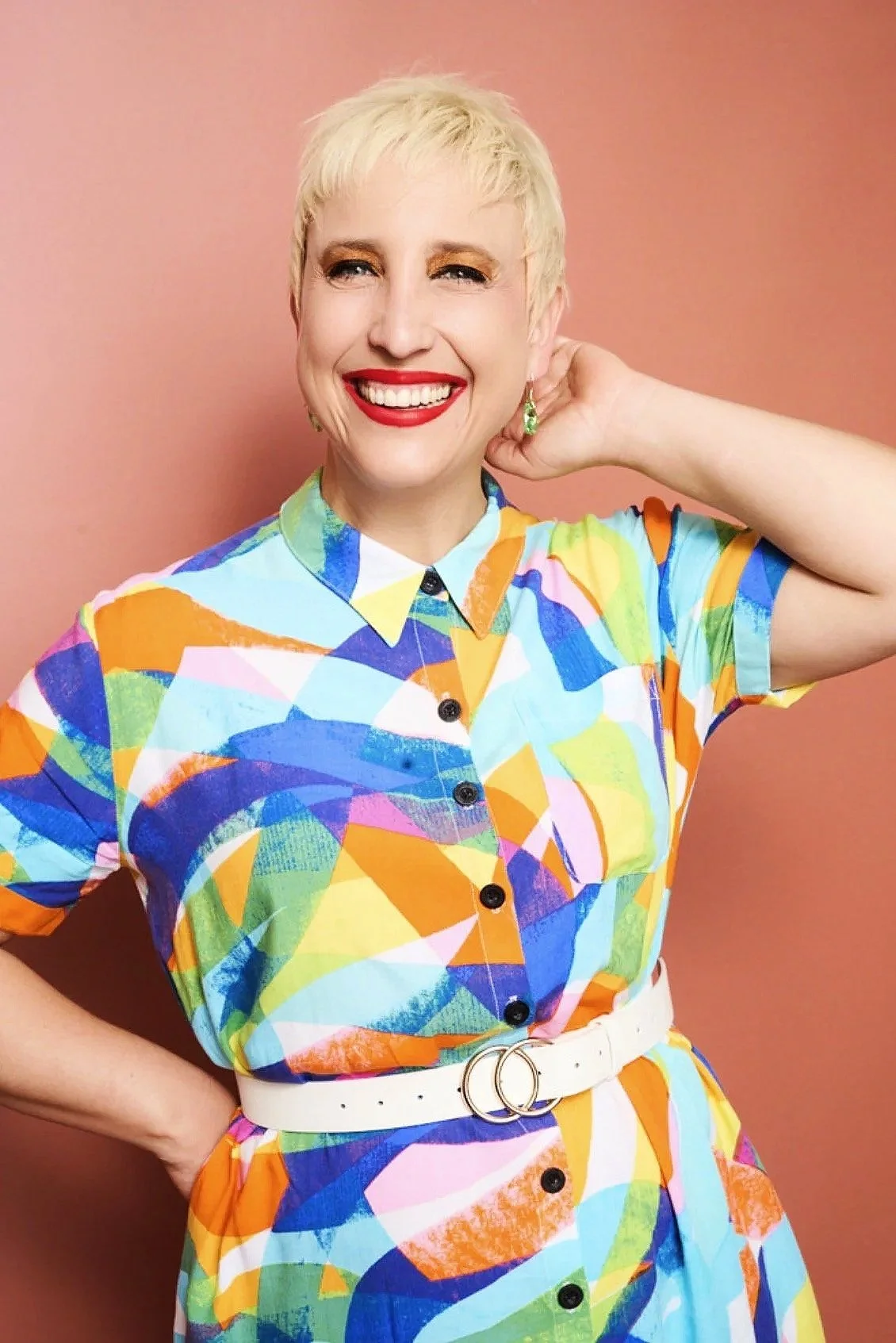 Woman with short platinum blonde hair smiling, wearing colorful geometric patterned dress with a white belt, standing against a pink background.