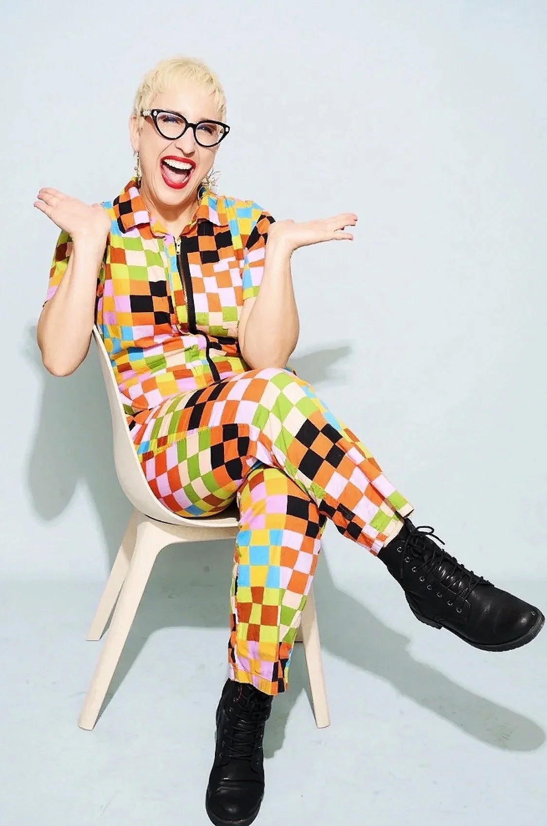A woman with short blonde hair, glasses, and red lipstick is sitting on a white chair against a plain white wall. She is wearing a colorful, checkered jumpsuit and black combat boots. She appears to be smiling and making a playful, expressive gesture with her hands.