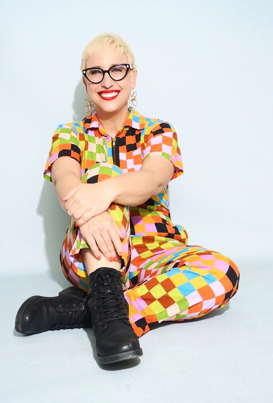 Woman sitting on the floor against a plain white background, wearing colorful checkered clothing, black boots, glasses, and earrings, smiling kindly.