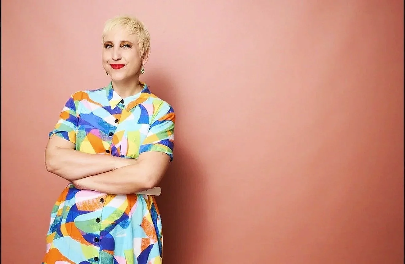A woman with short blonde hair standing with arms crossed, wearing a colorful abstract patterned dress, in front of a pink wall.