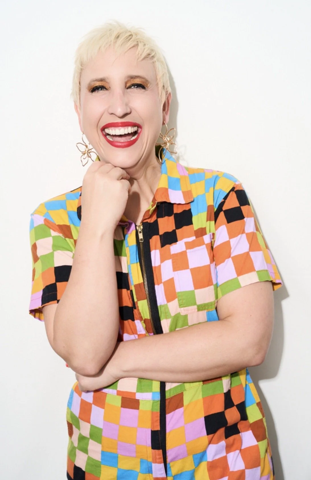 A woman with short blonde hair, red lipstick, and colorful checkered clothing, smiling with her hand under her chin against a plain white background.