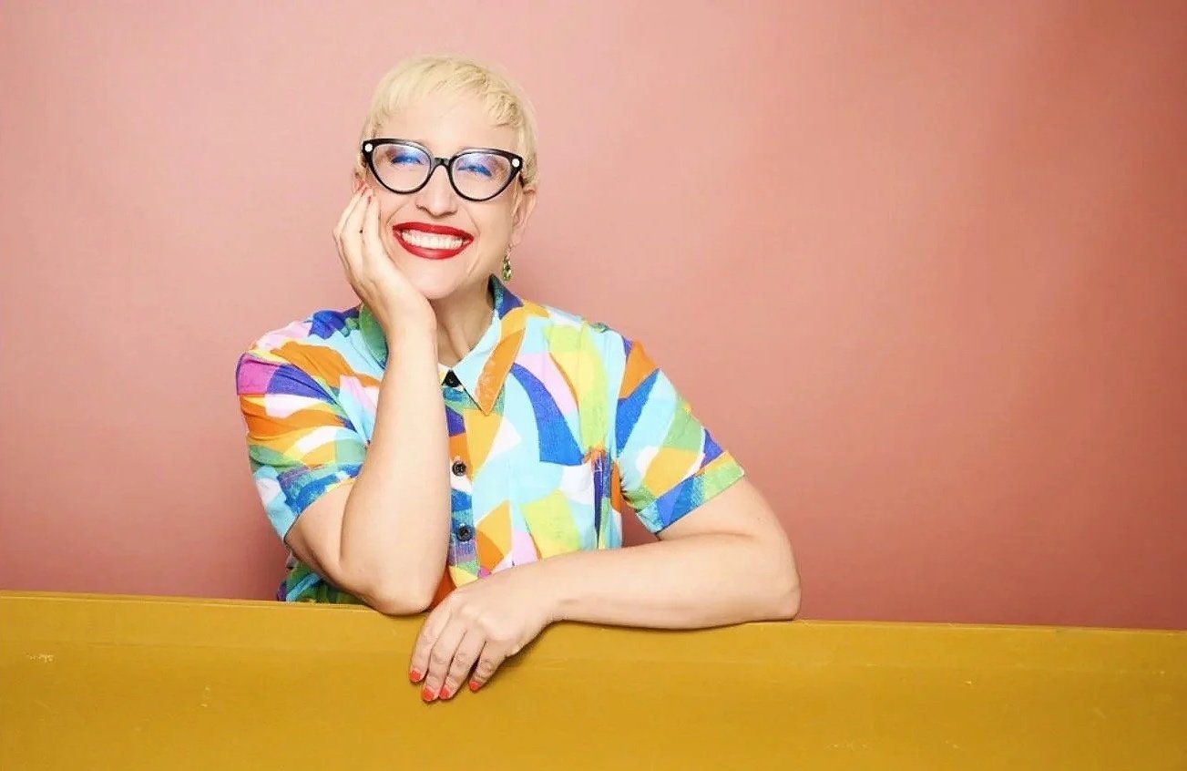 A woman with short blonde hair, glasses, and bright red lipstick smiling and resting her face on her hand while sitting at a yellow table against a pink background. She is wearing a colorful, abstract patterned short-sleeve shirt.