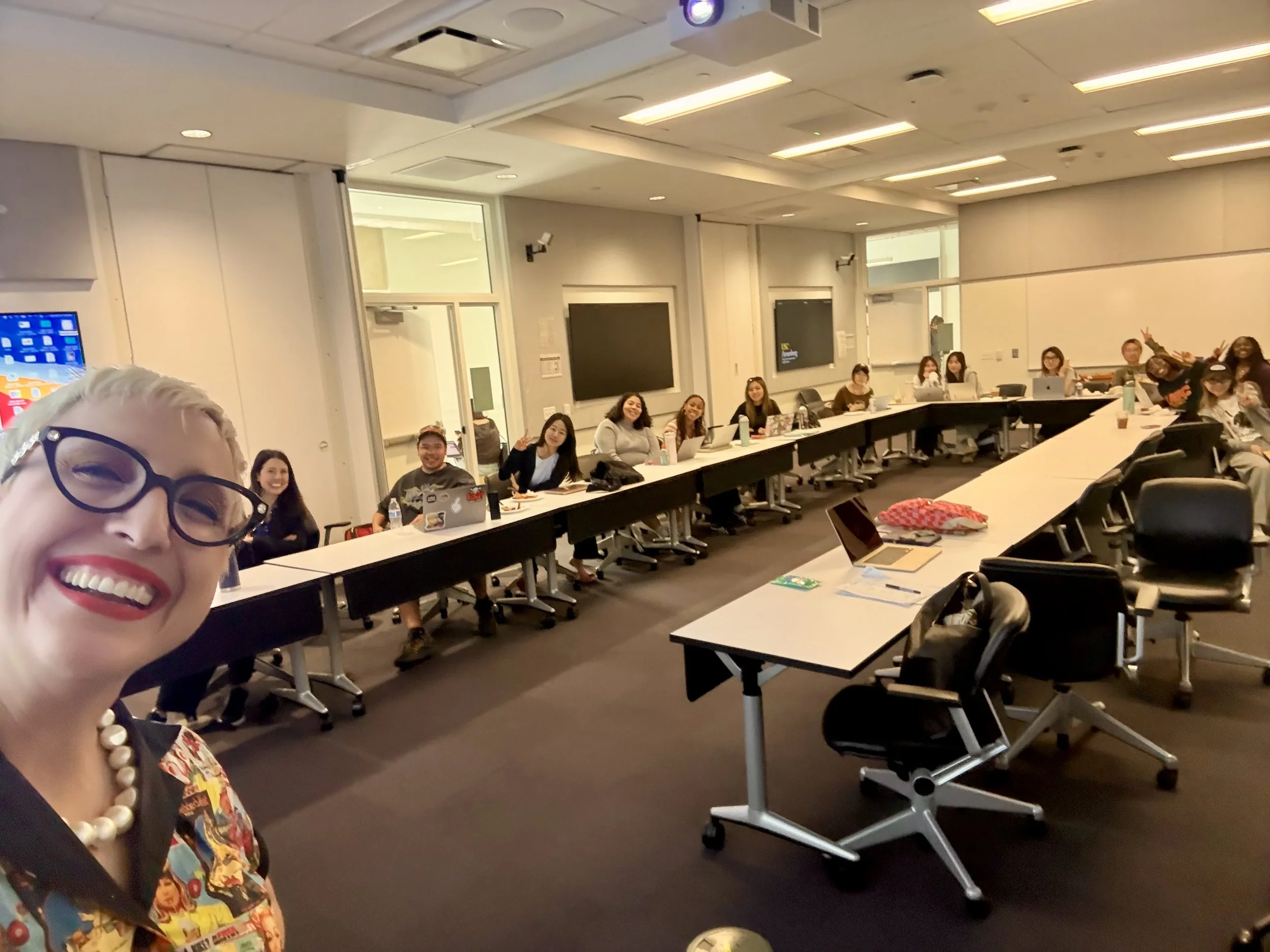A woman with glasses and colorful clothing taking a selfie in a conference room with about 12 people seated at long tables, smiling and looking at the camera. The room has white walls, windows, and multiple large screens on the wall.
