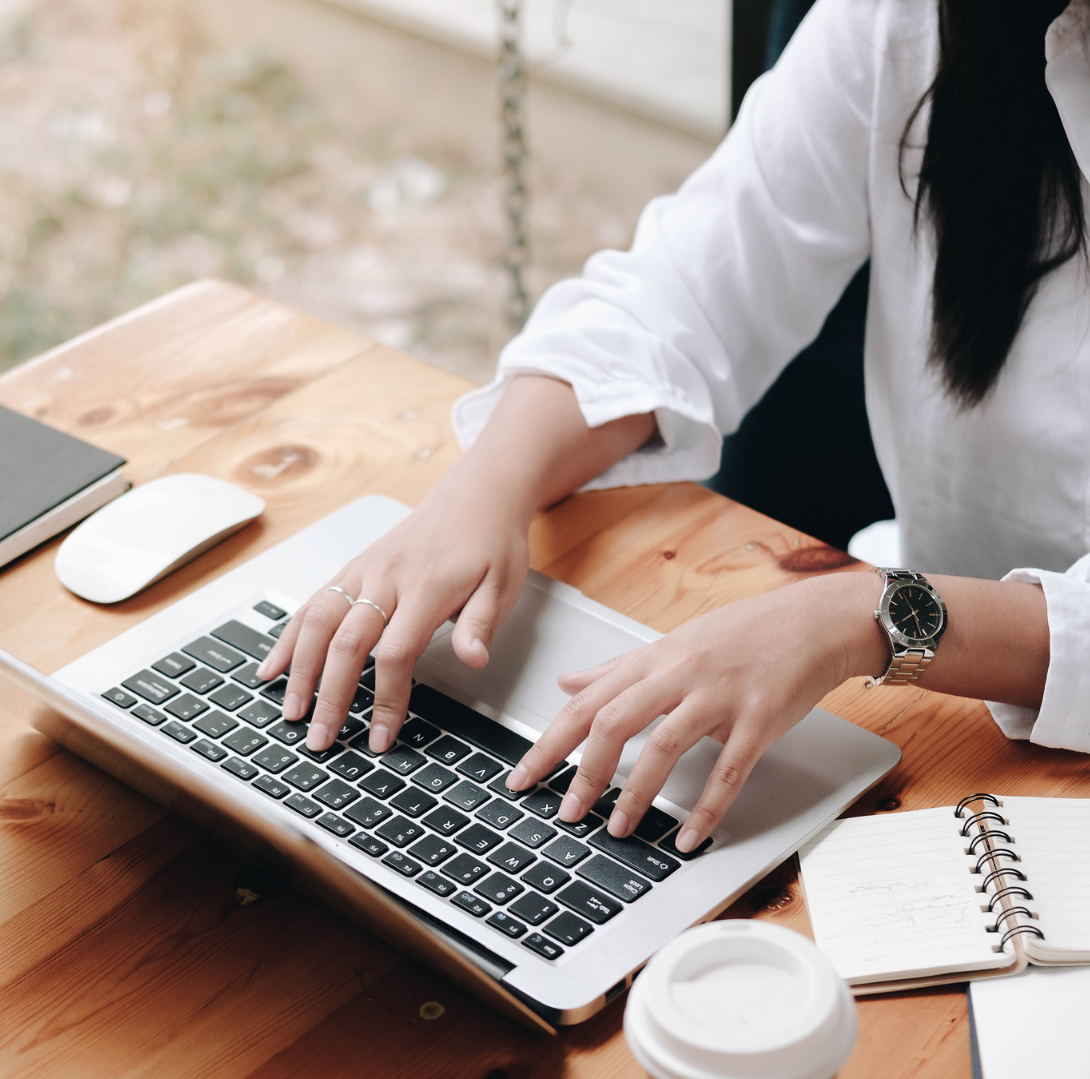 A woman typing on a silver laptop with a black keyboard, wearing a white shirt and a silver wristwatch, sitting at a wooden desk near a window.