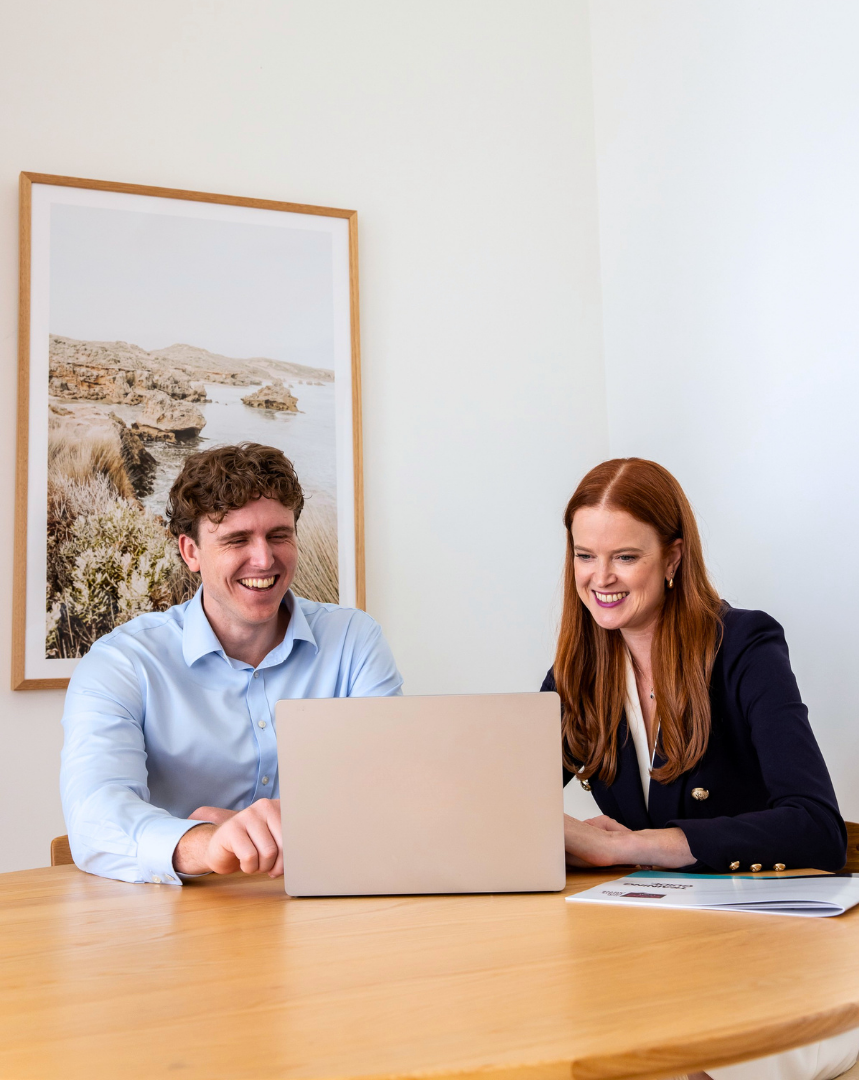 Two people, a man and a woman, sitting at a desk and smiling while looking at a laptop screen in a bright office with a framed nature photograph on the wall.