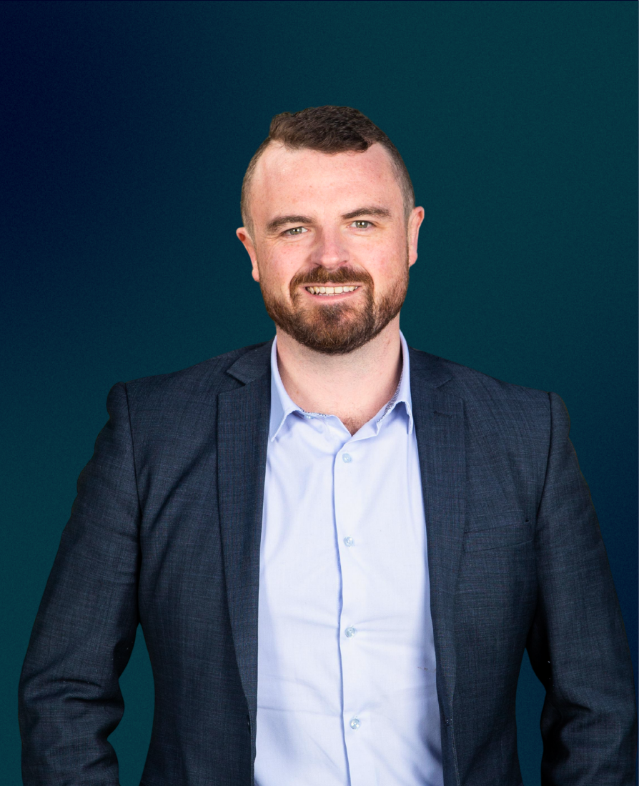 A man with short brown hair and a beard, wearing a navy blazer and a light blue dress shirt, smiling at the camera against a dark green background.