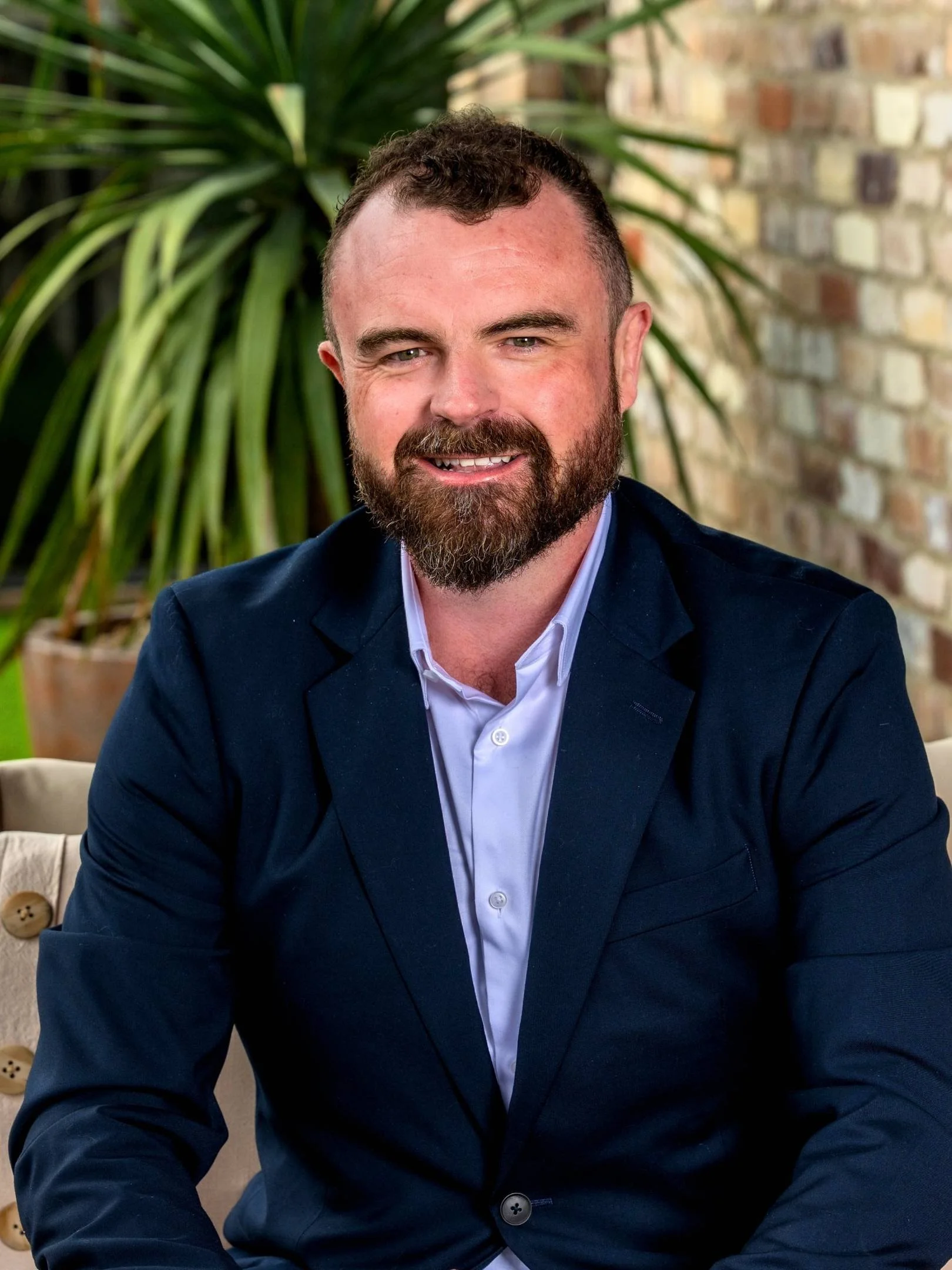 A man in a dark blue suit and white shirt sitting in front of a stone fireplace, smiling at the camera.