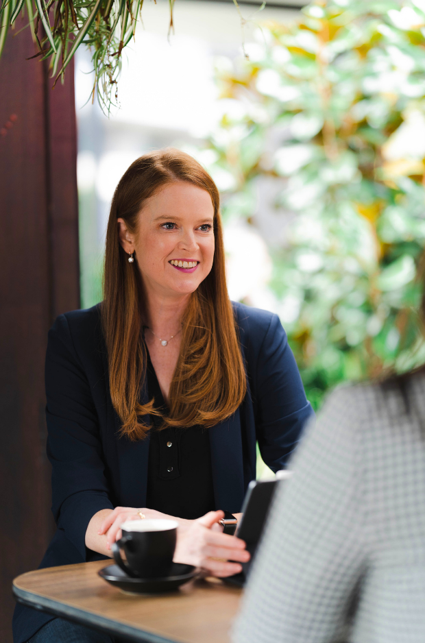 A woman with long red hair sitting at a table in a cafe, talking to another person, with a cup and saucer in front of her.