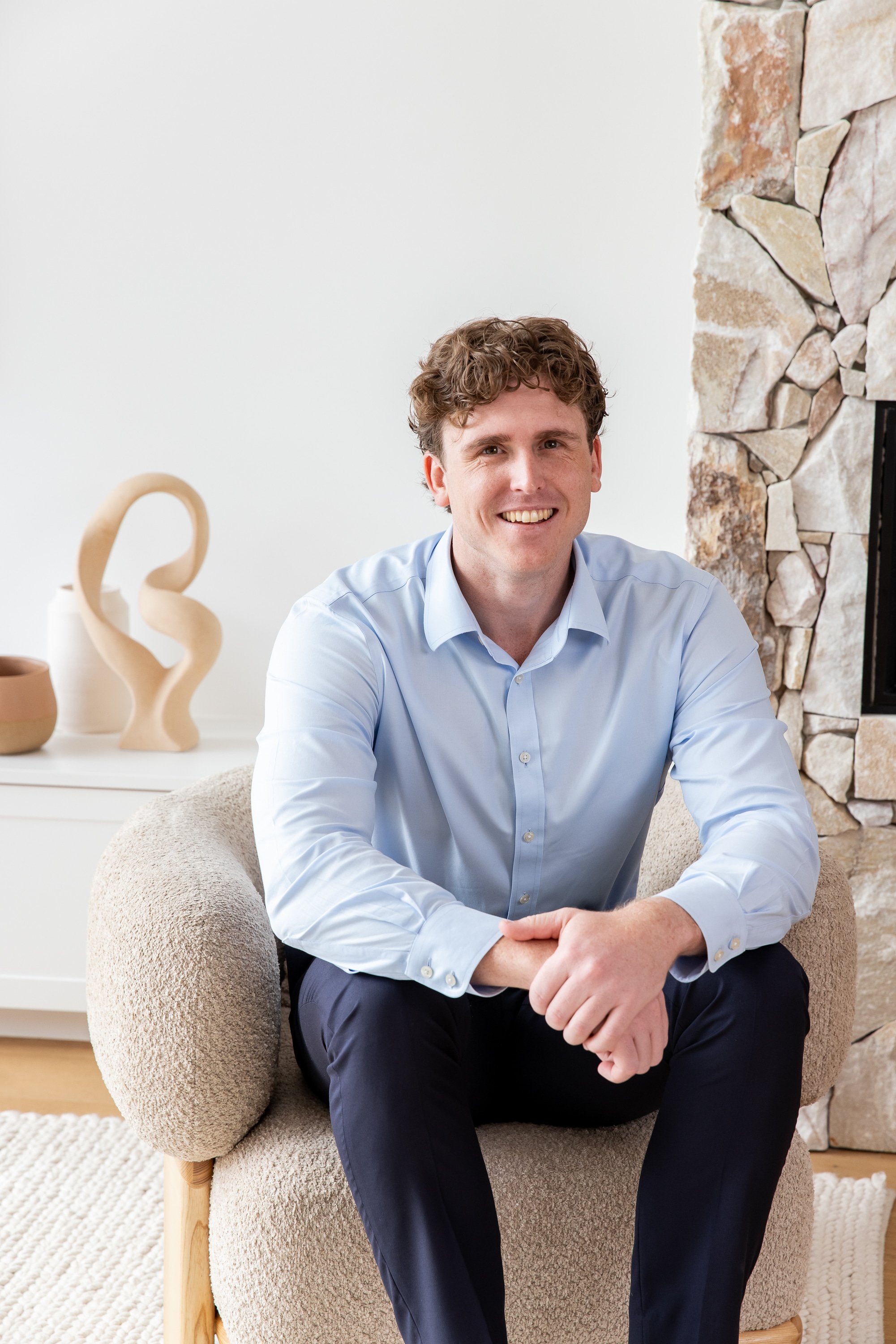 A young man in a light blue shirt and dark pants sitting on a beige armchair, smiling, with a stone fireplace and modern decorative art on the side in a bright living room.