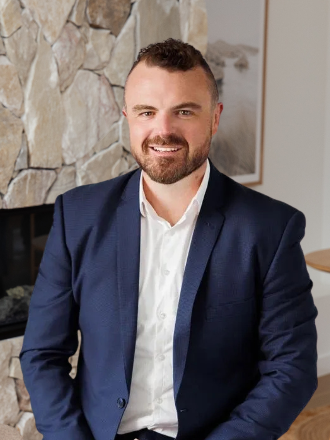A man in a dark blue suit and white shirt sitting in front of a stone fireplace, smiling at the camera.