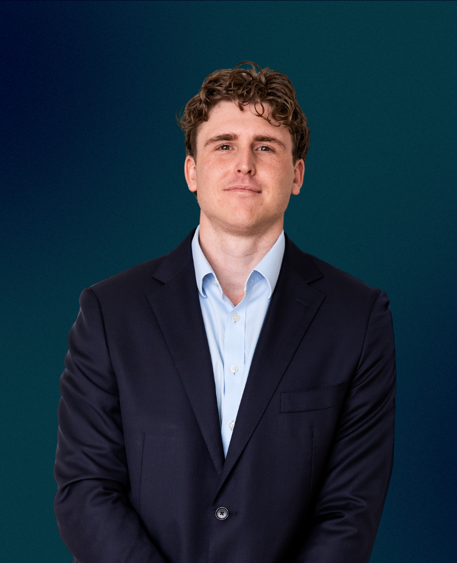 A young man with curly brown hair wearing a dark suit jacket and light blue dress shirt, standing against a teal background.
