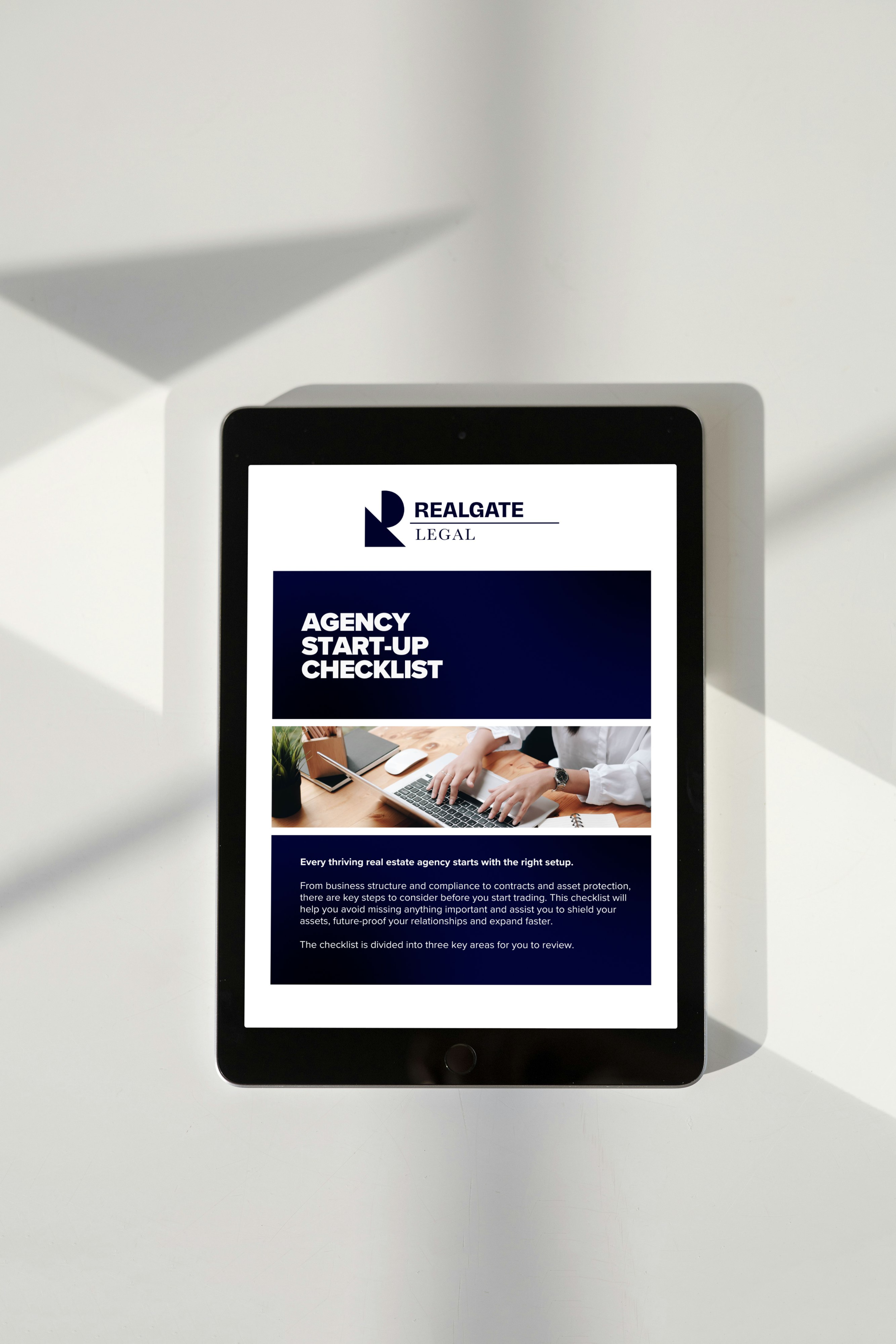 A tablet displaying a real estate agency start-up checklist from RealGATE Legal on a white surface with a shadow cast over the top left corner.