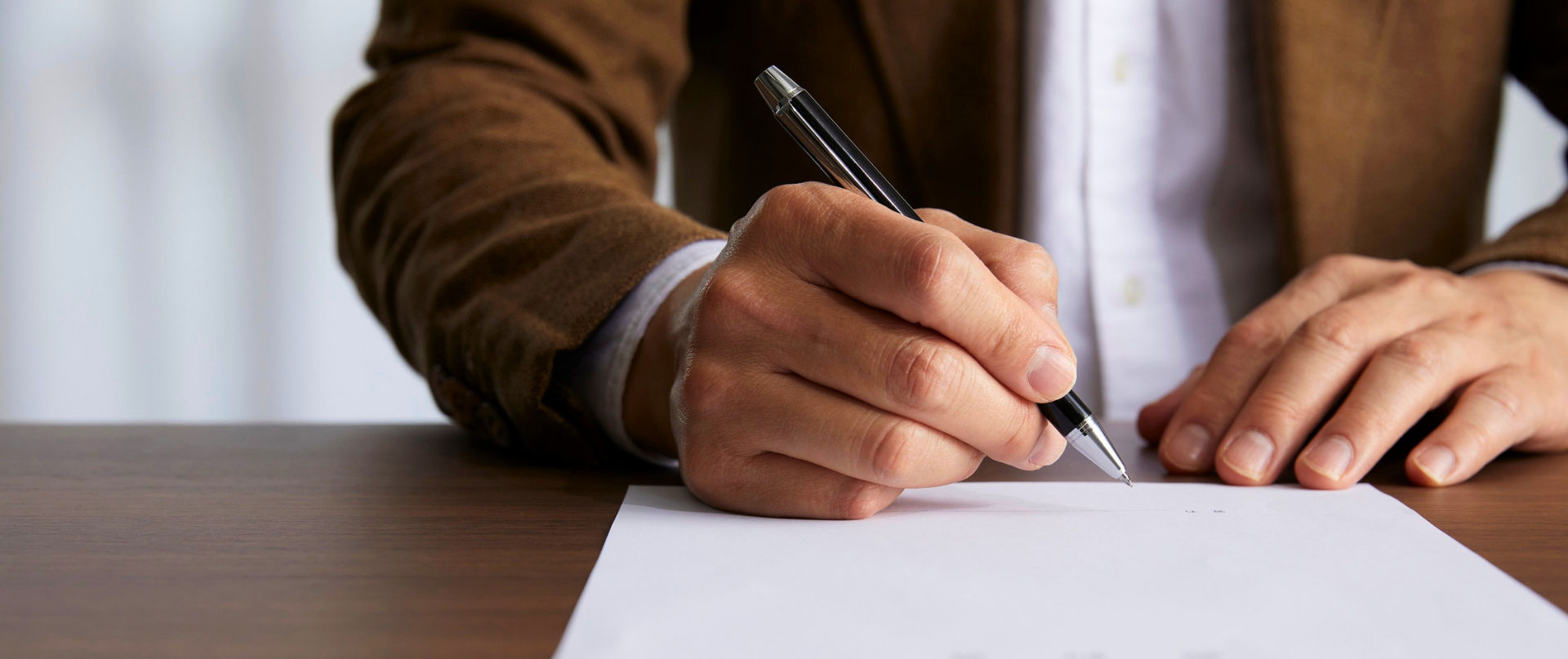 A person wearing a brown jacket and white shirt is writing on a blank sheet of paper with a black pen on a wooden surface.