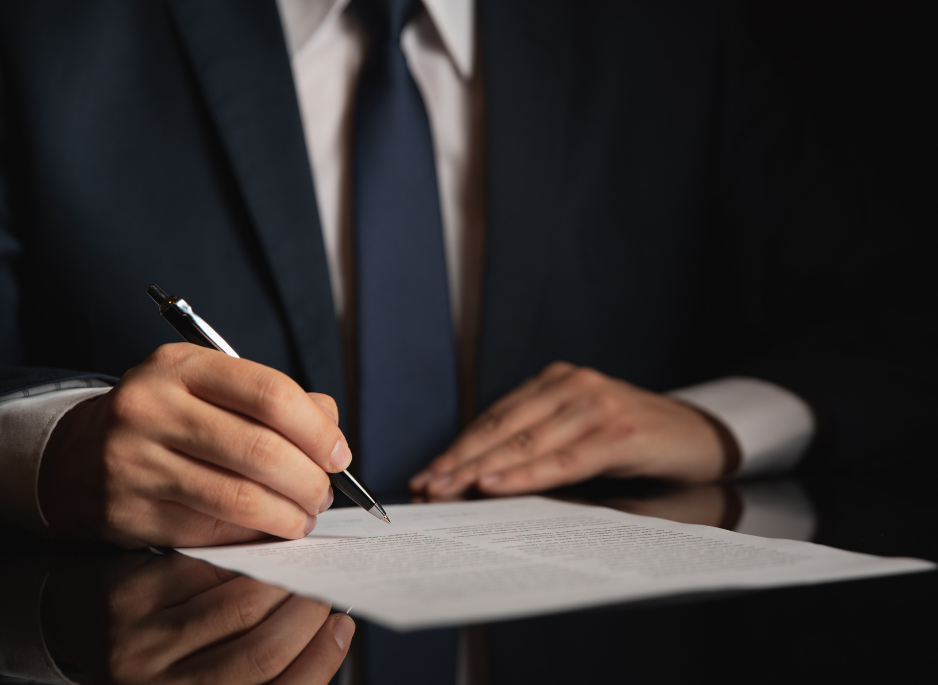 A person in a suit signing a document with a black pen on a dark reflective surface.