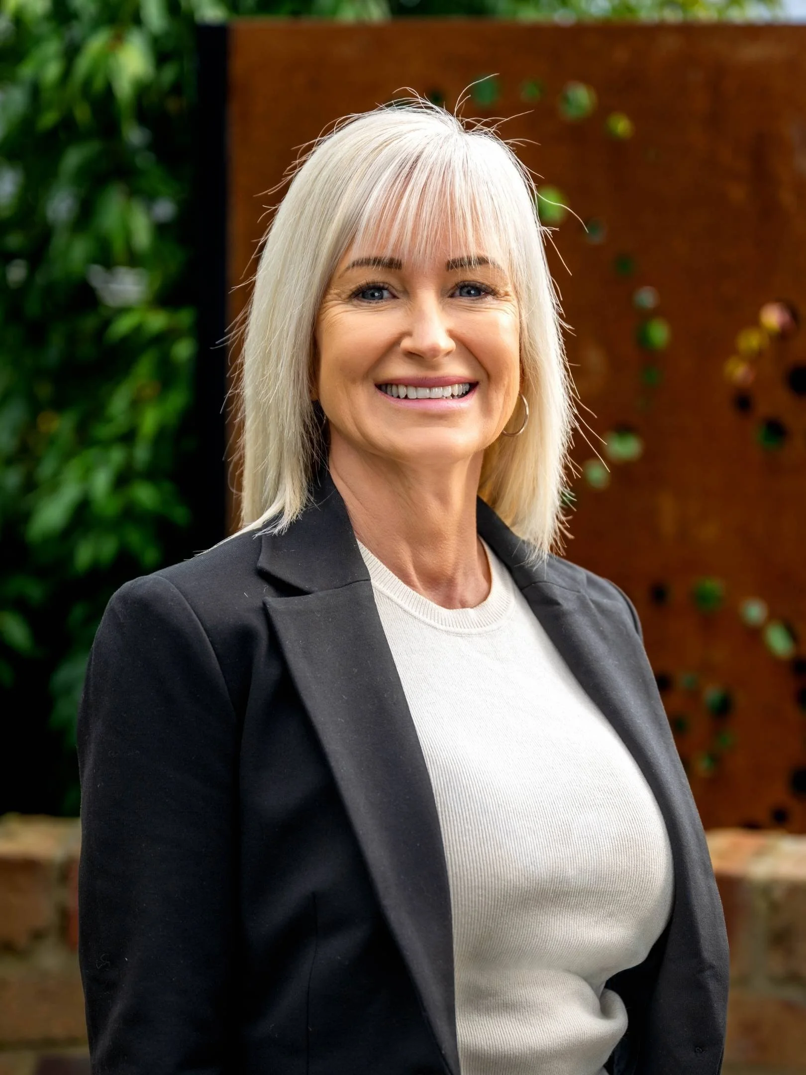 A middle-aged woman with blonde hair and blue eyes wearing a black turtleneck and blazer, standing in a room with a stone wall and plant in the background.