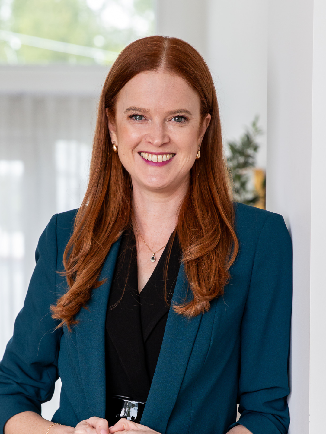 A woman with red hair and blue eyes smiling, wearing a navy blazer over a black top, gold earrings, a necklace, and a belt, standing indoors with a blurred background of a window and plants.