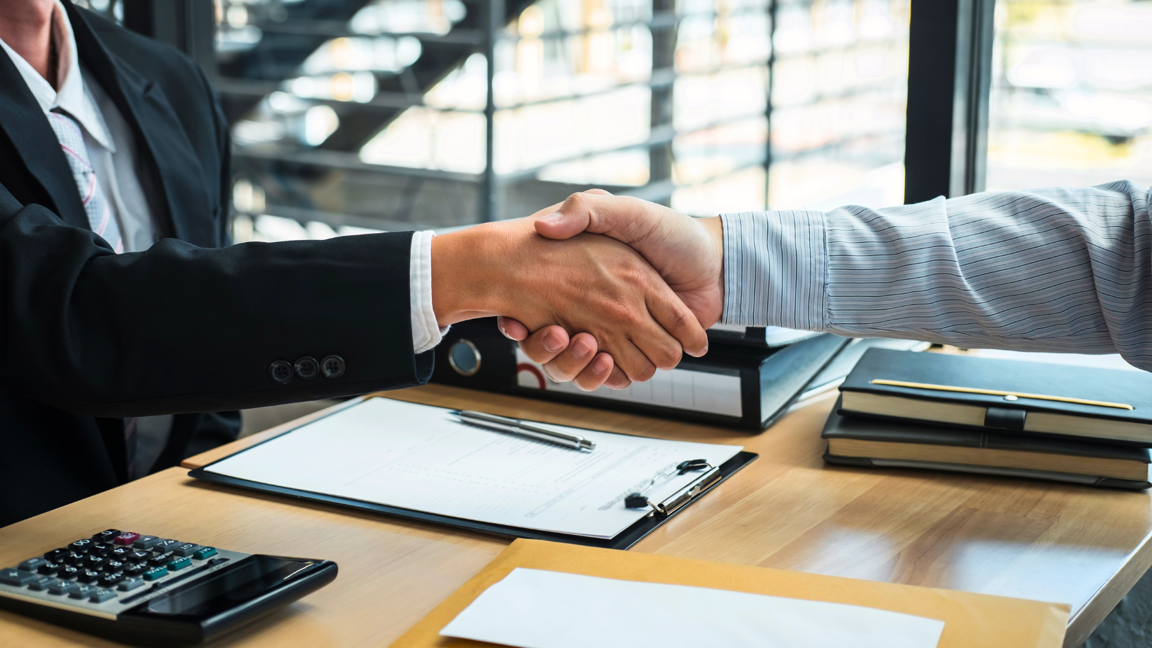 Two individuals shaking hands across a desk in a professional setting, with documents, a calculator, and files on the desk.