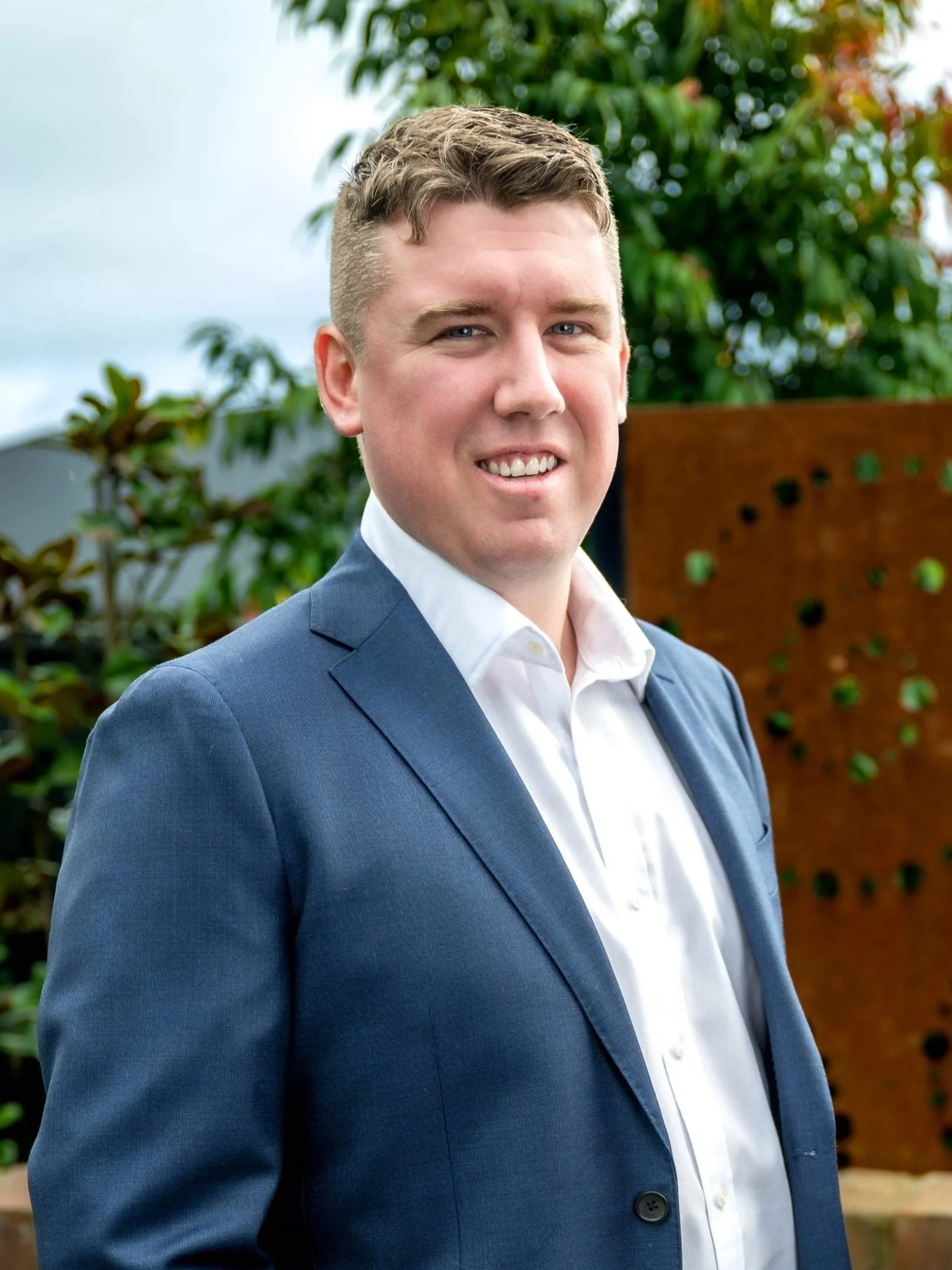 A young man with light brown hair and blue eyes in a dark blue plaid suit, white shirt, and navy polka dot tie, smiling confidently in a bright room with large windows and sheer curtains.