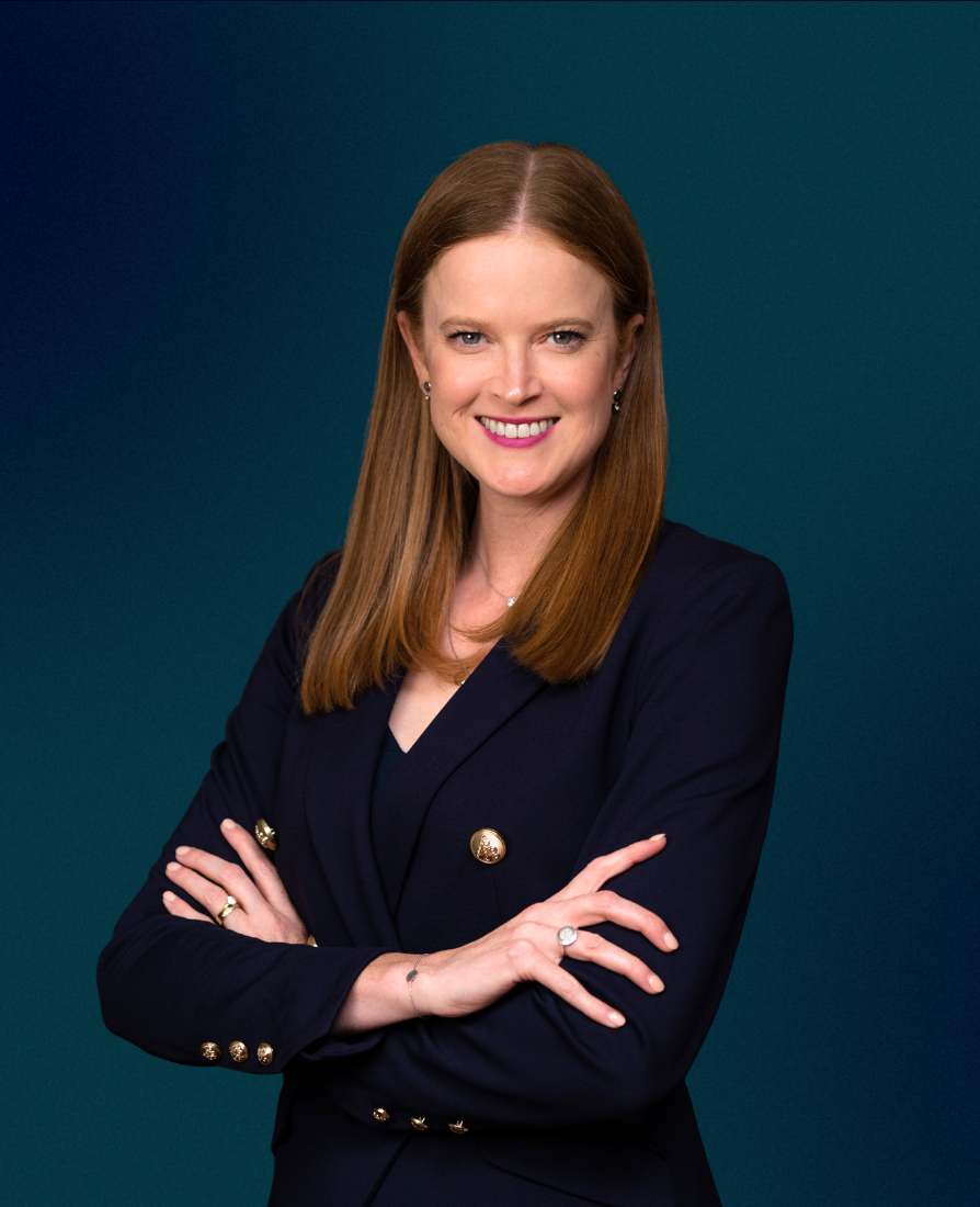 A woman with straight red hair, wearing a navy blazer with gold buttons, stands with arms crossed, smiling against a teal background.