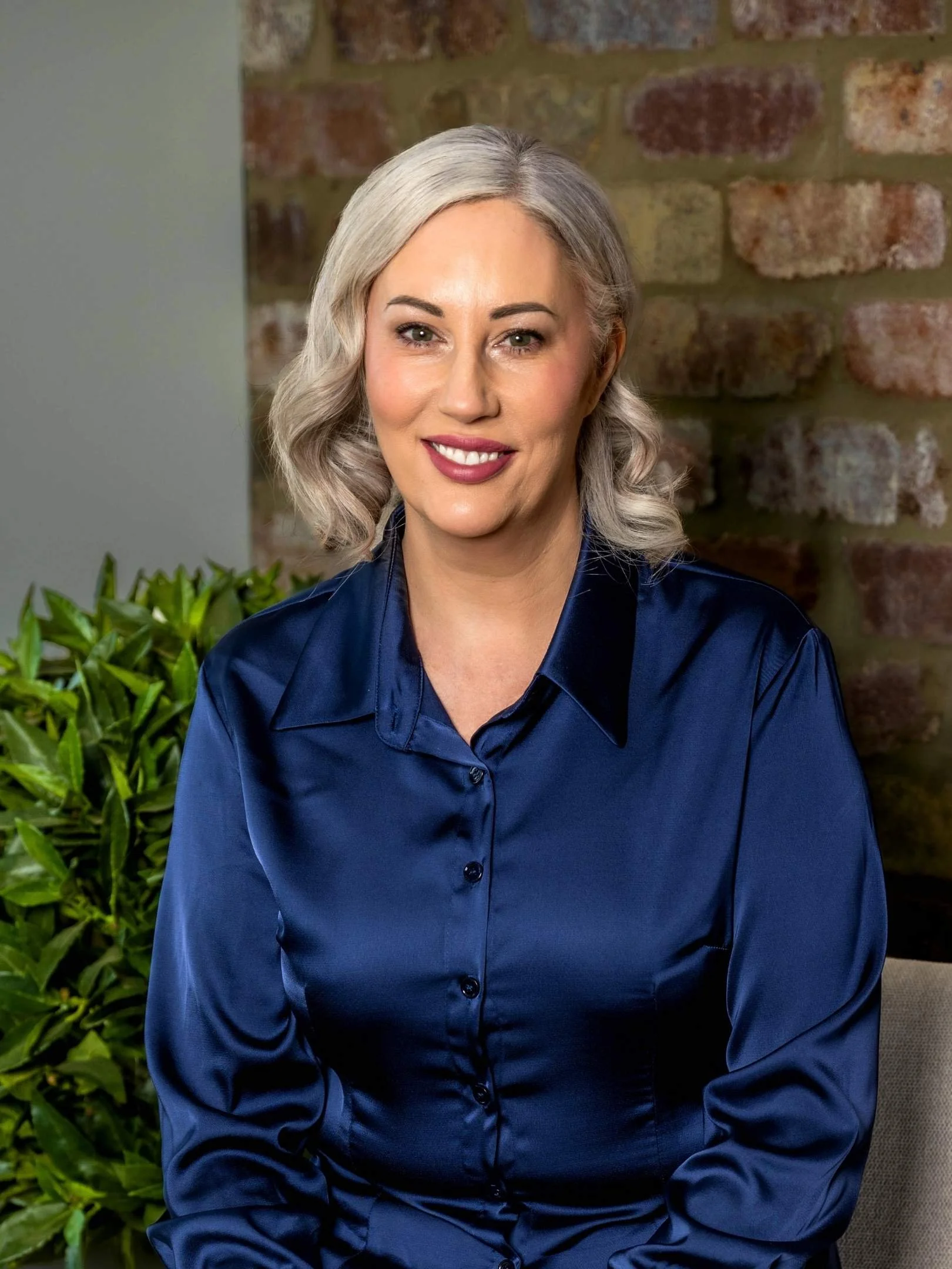 A woman with blonde hair styled in loose waves, wearing a navy blazer and white top, smiling, standing in front of a stone wall.