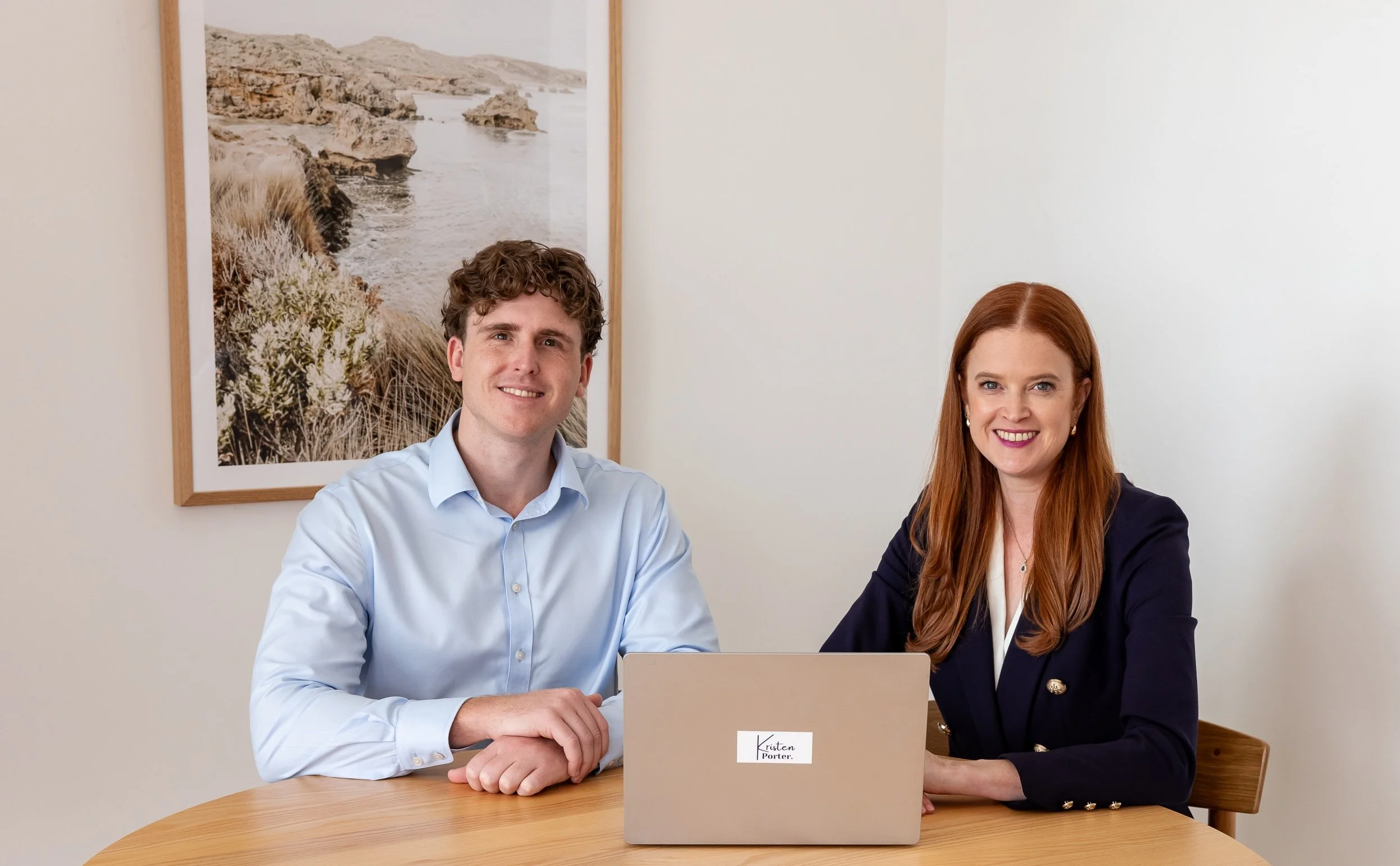 A woman with long red hair and a navy blazer sitting at a wooden table with a laptop and a man with curly brown hair wearing a light blue dress shirt. Behind them is a framed landscape photo on a light wall.