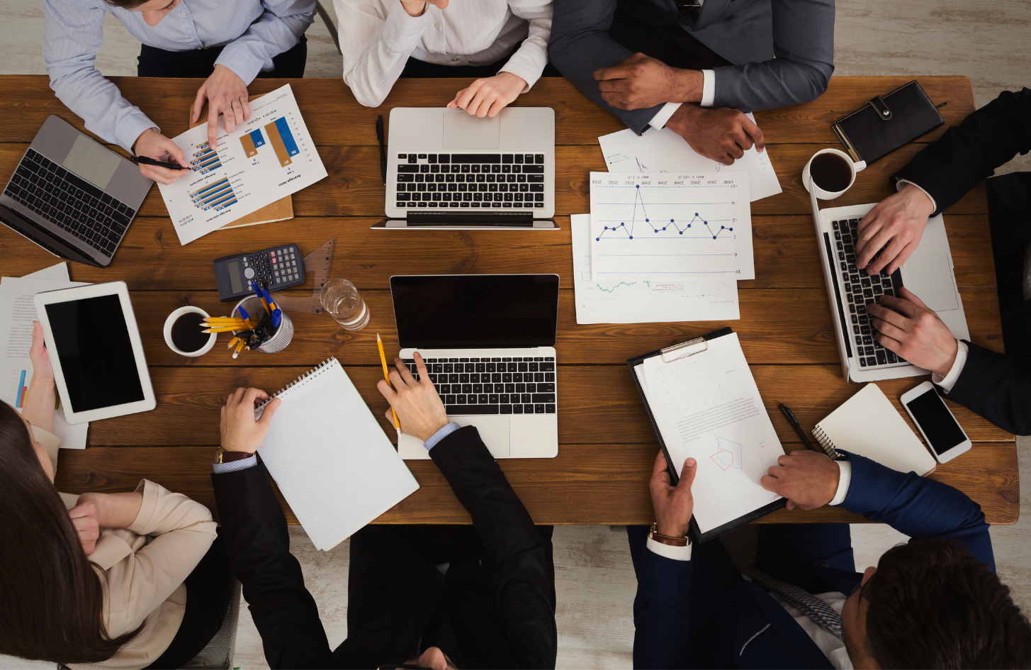 Top-down view of six people working at a wooden table with laptops, tablets, documents, charts, graphs, and office supplies in a business meeting.