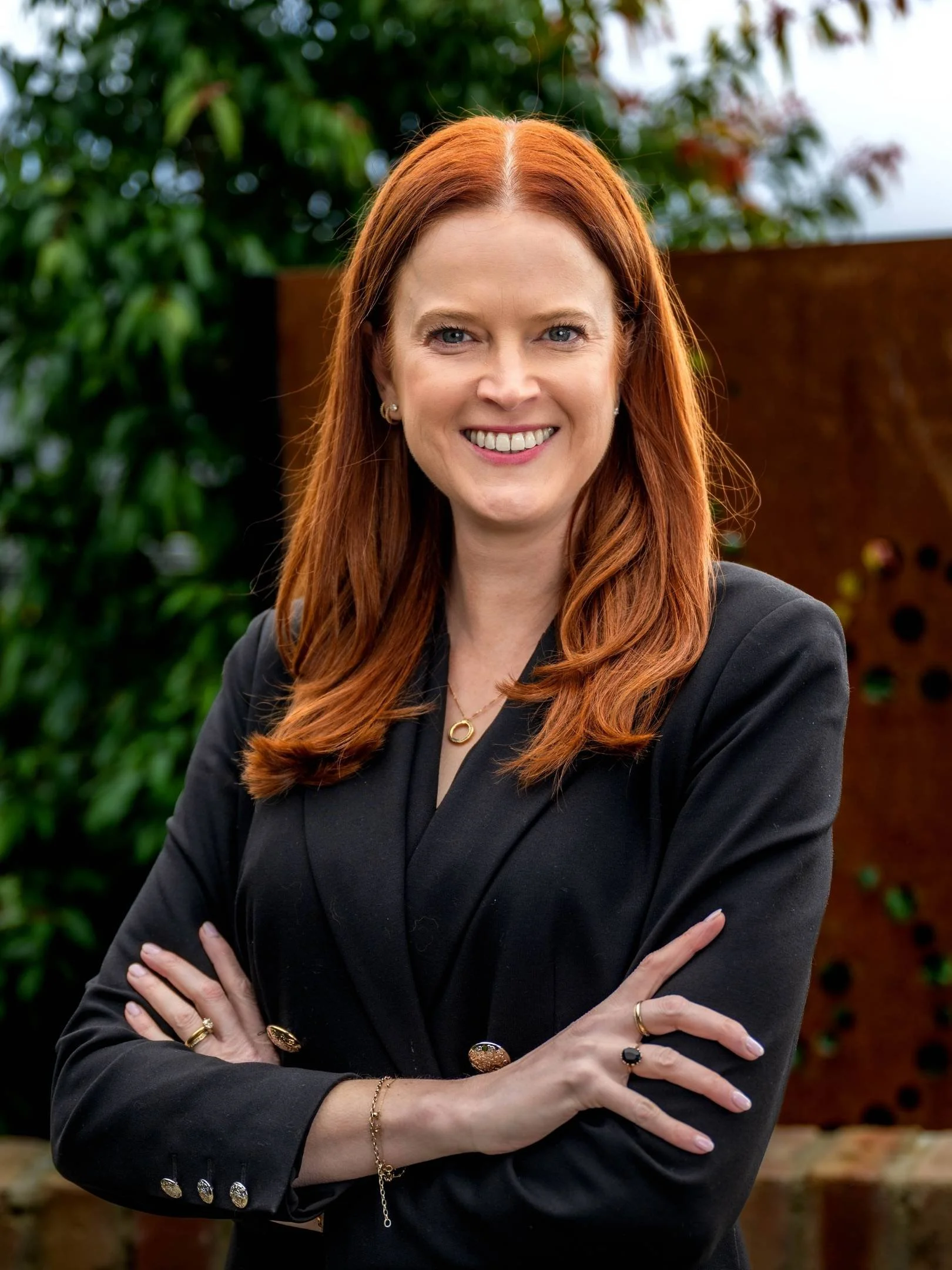 A woman with red hair and blue eyes smiling, wearing a navy blazer over a black top, gold earrings, a necklace, and a belt, standing indoors with a blurred background of a window and plants.