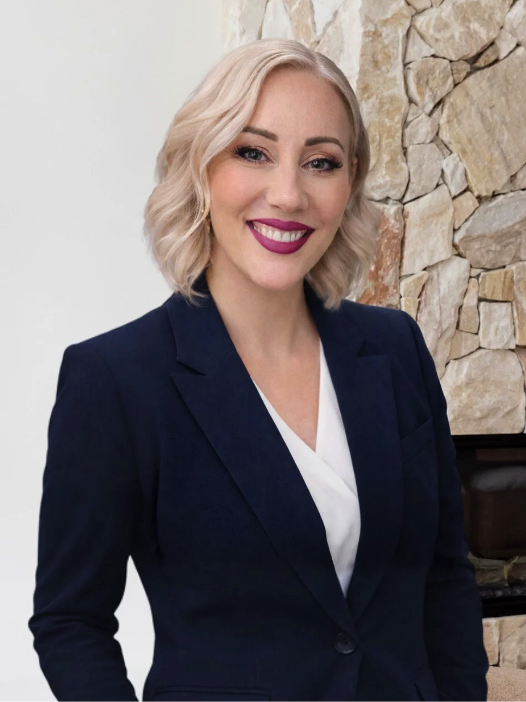 A woman with blonde hair styled in loose waves, wearing a navy blazer and white top, smiling, standing in front of a stone wall.