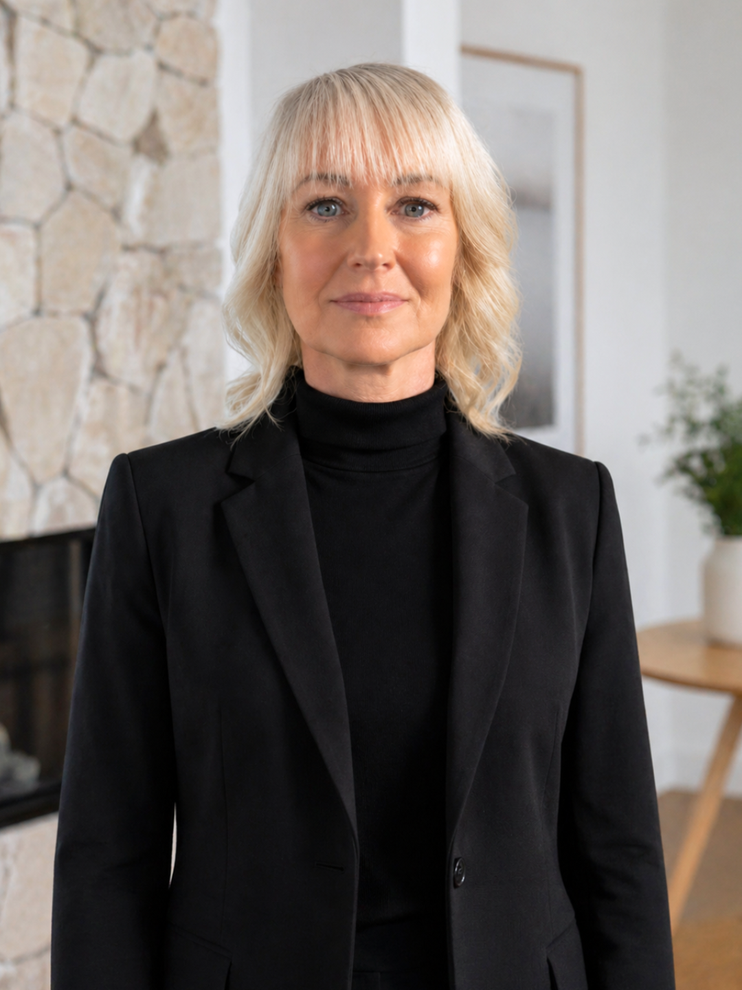 A middle-aged woman with blonde hair and blue eyes wearing a black turtleneck and blazer, standing in a room with a stone wall and plant in the background.