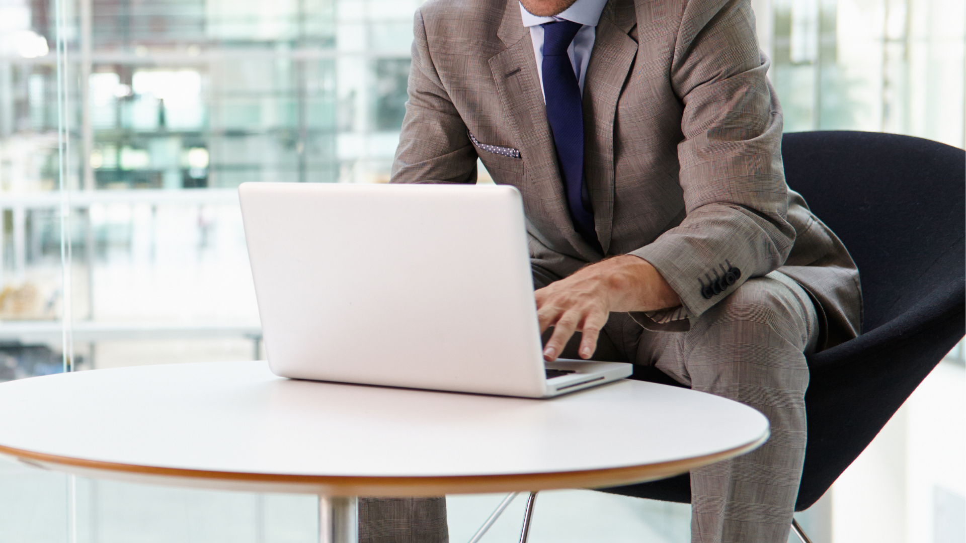 Businessman in a gray suit working on a laptop at a white table in a modern office.
