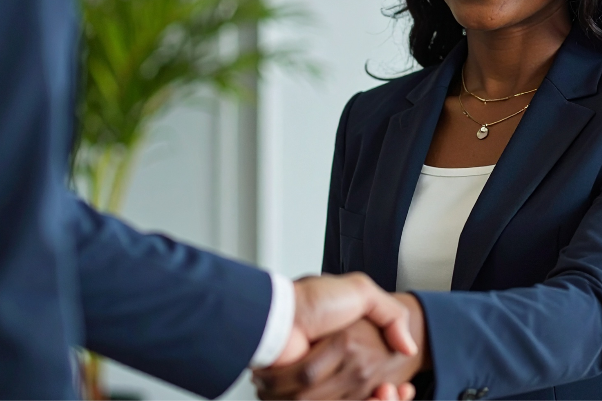 Close-up of a professional handshake between two individuals, one woman wearing a dark blazer and a white blouse, with layered necklaces, in a bright office setting with a plant in the background.