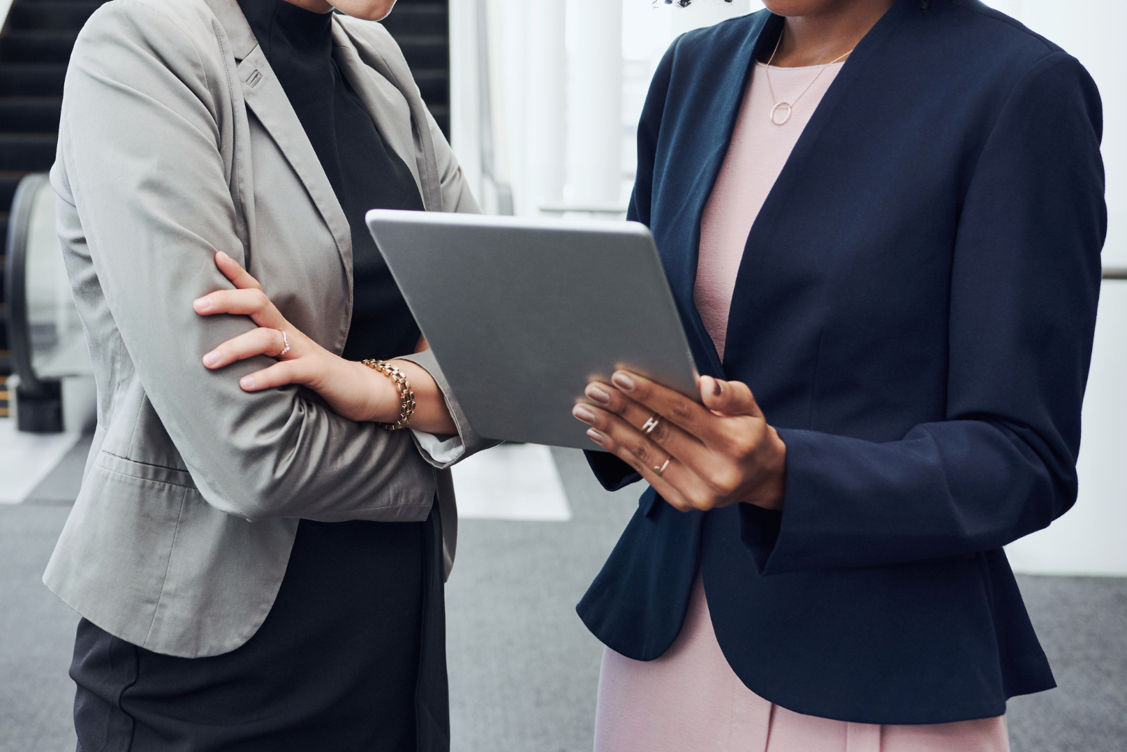 Two women in business attire standing indoors, looking at a tablet.
