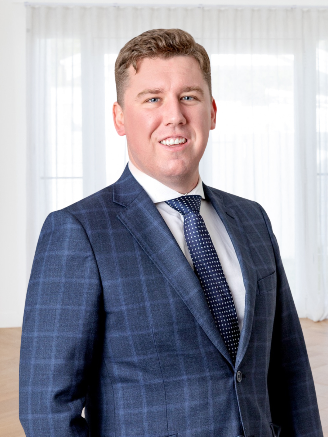 A young man with light brown hair and blue eyes in a dark blue plaid suit, white shirt, and navy polka dot tie, smiling confidently in a bright room with large windows and sheer curtains.