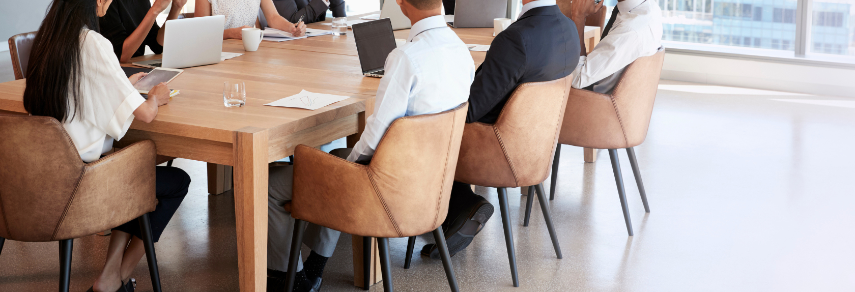 Business meeting with several people seated around a wooden conference table with laptops, notebooks, and coffee cups, in a modern office space with large windows.