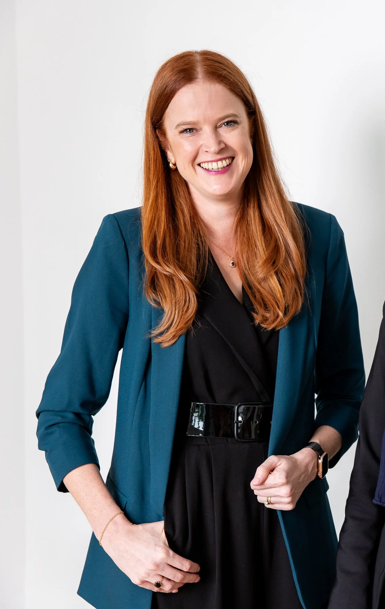 A woman with long red hair, smiling, wearing a teal blazer over a black top with a wide black belt, standing against a plain white background.