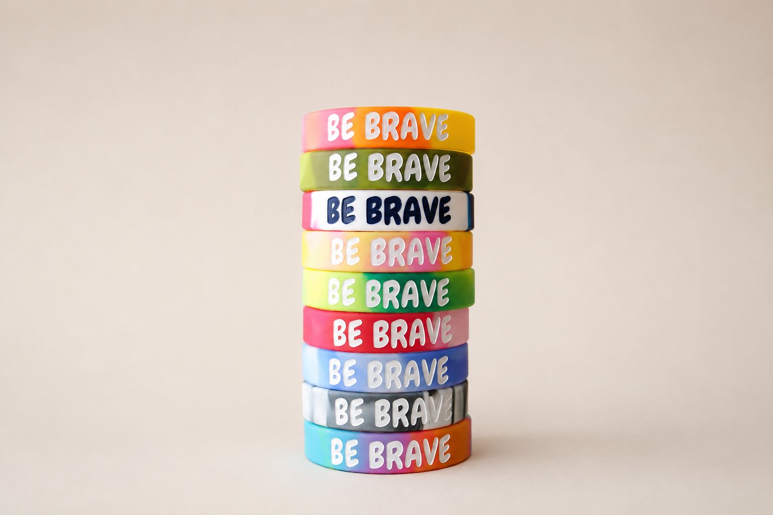 Stack of colorful wristbands with the phrase 'BE BRAVE' on each band, in various colors including pink, yellow, green, navy, orange, white, red, blue, gray, purple, and multicolor, arranged vertically on a plain beige background.