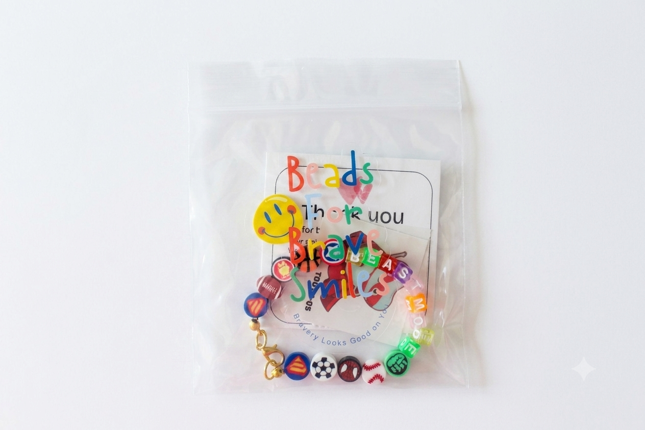 Colorful beaded bracelet with sports and superhero themed charms, enclosed in a clear plastic bag with a note saying 'Beads, Thank you, for, love, smiles' on a white background.