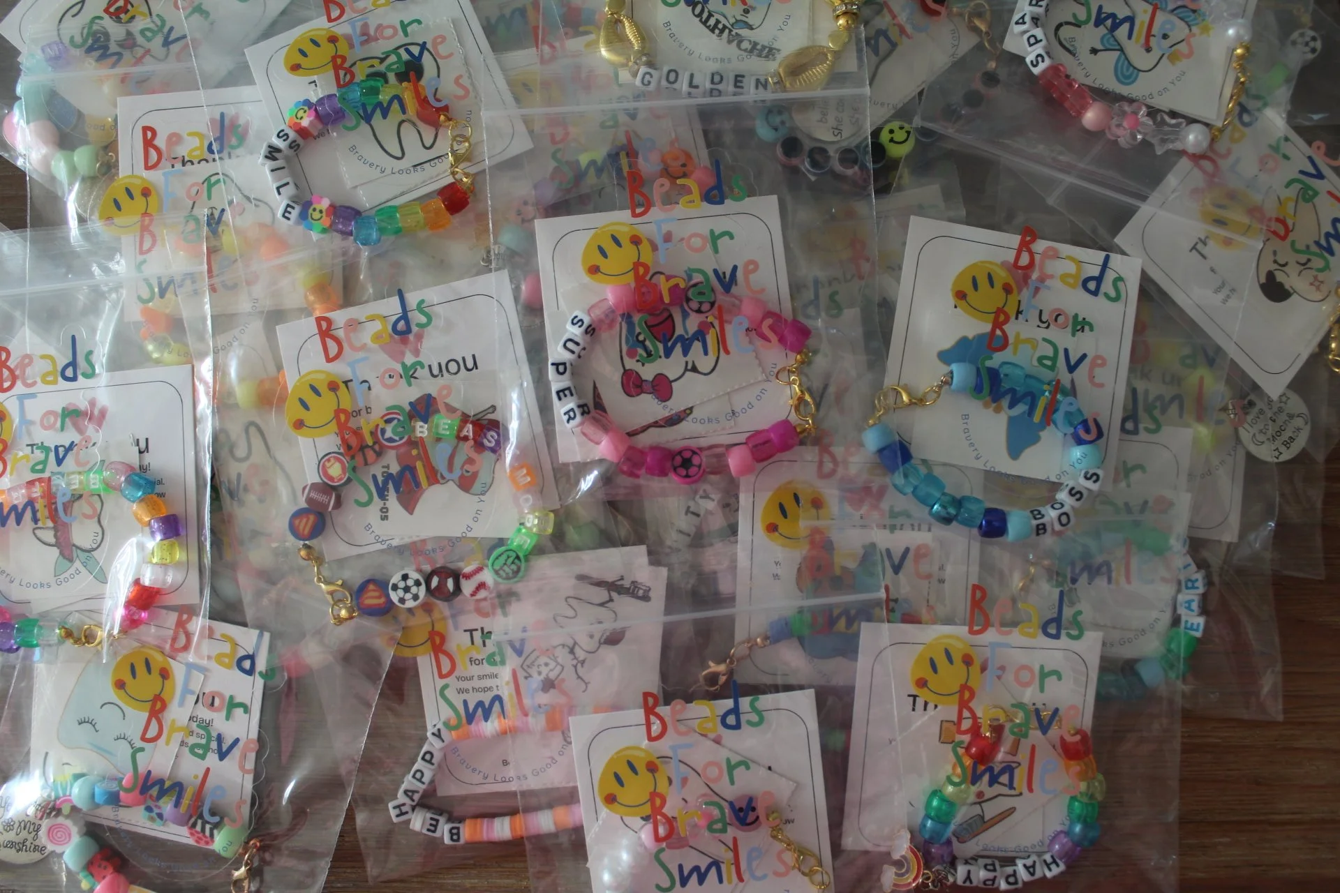 Multiple packages of colorful beaded bracelets with various bead designs, in clear plastic bags, each featuring a 'Beads for Bravery Smiles' logo with a yellow smiley face, displayed on a wooden surface.