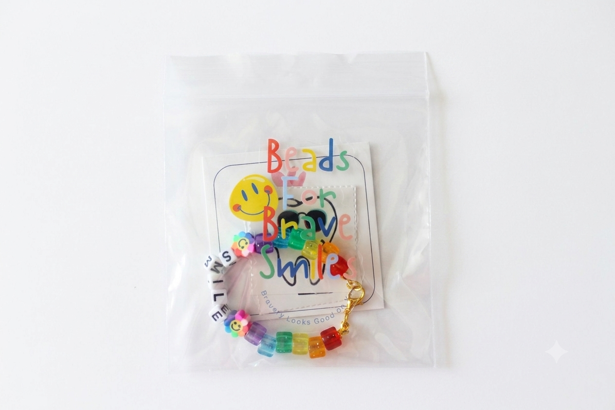 Colorful beaded bracelet with smiley face charms, inside clear plastic packaging labeled "Beads For Brave Smiles," against a white background.