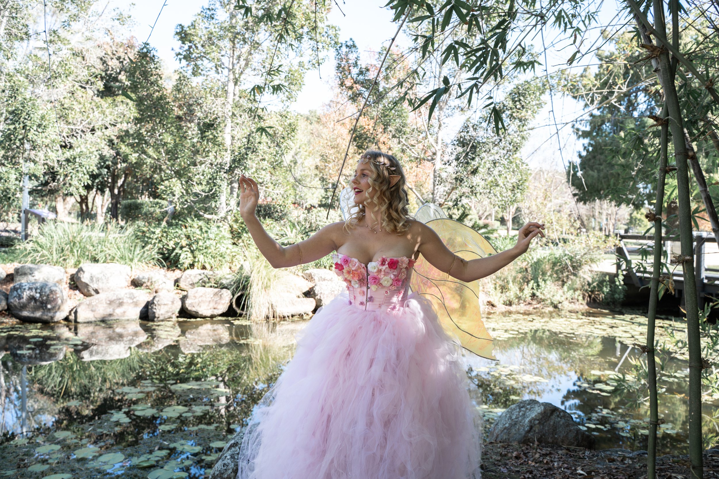A woman dressed in a pink fairy or princess costume with fairy wings, standing near a pond in a garden, surrounded by trees and rocks.