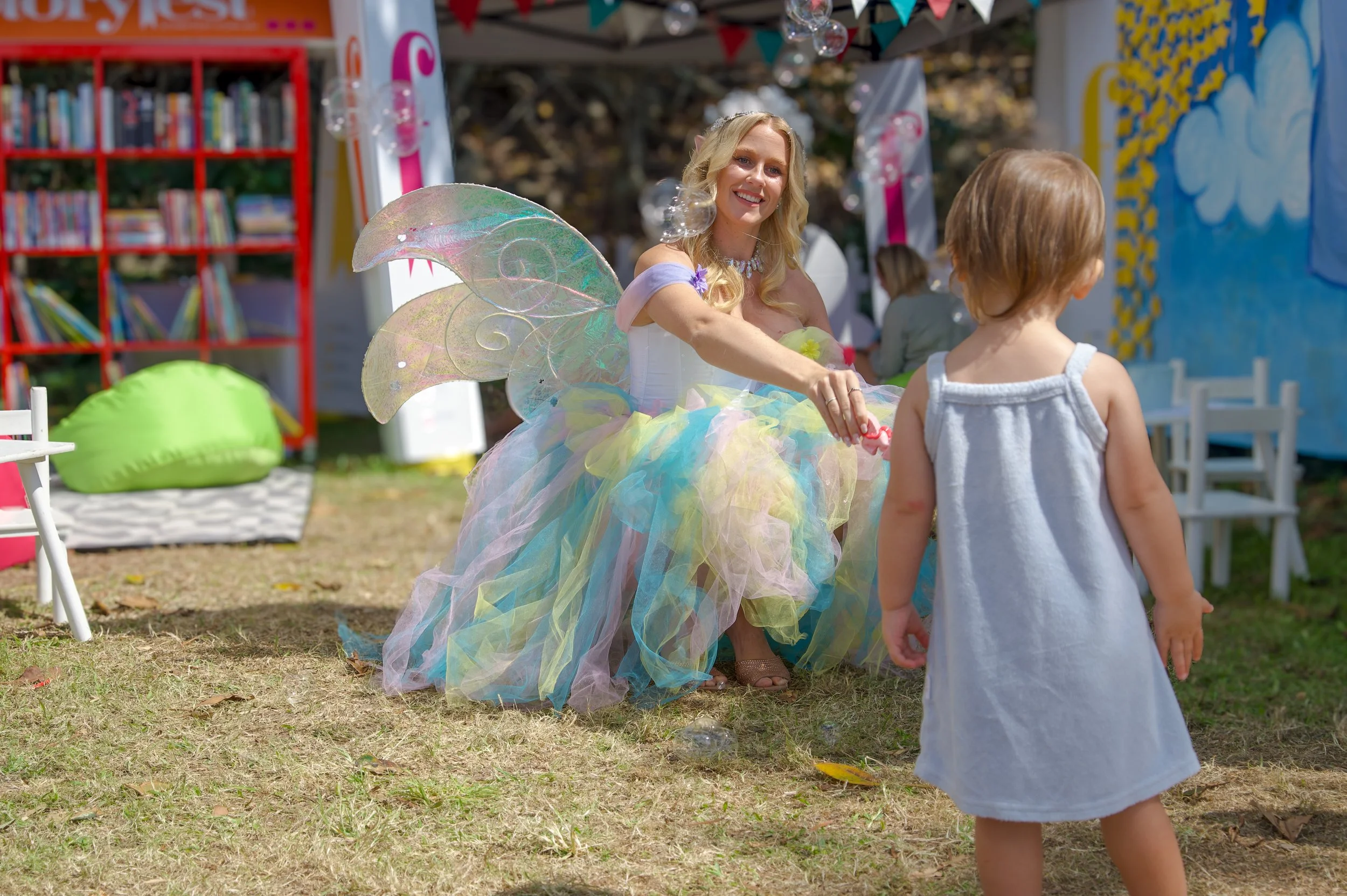 A woman dressed as a fairy with iridescent wings and a colorful tutu interacts with a young girl at an outdoor event or party, surrounded by decorations and bookshelves.