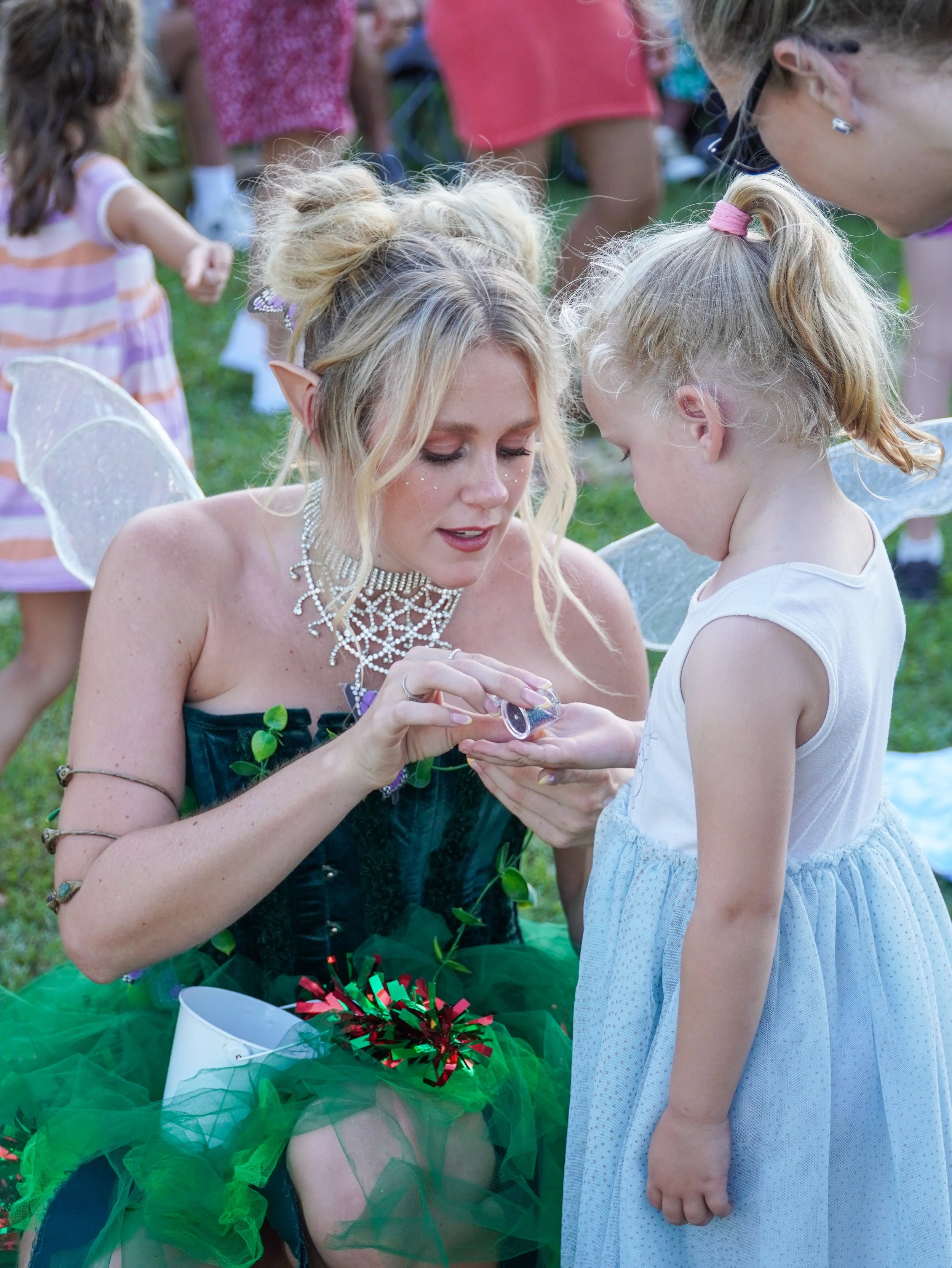 A woman dressed as a fairy or fantasy character, wearing a green costume, fairy wings, and a green tulle skirt, is holding a small container with glitter or confetti. She is focused on a young girl with blonde hair in a ponytail, wearing a sleeveless white dress. The scene appears to be at an outdoor event with other children and people in the background.