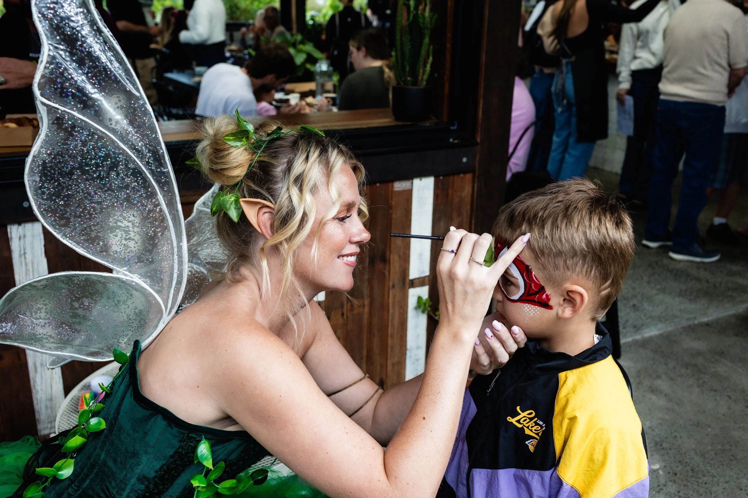 A woman dressed as a fairy with sparkly wings, a headband with green leaves, and green leaf accents on her dress, paints the face of a young boy at a busy outdoor event.