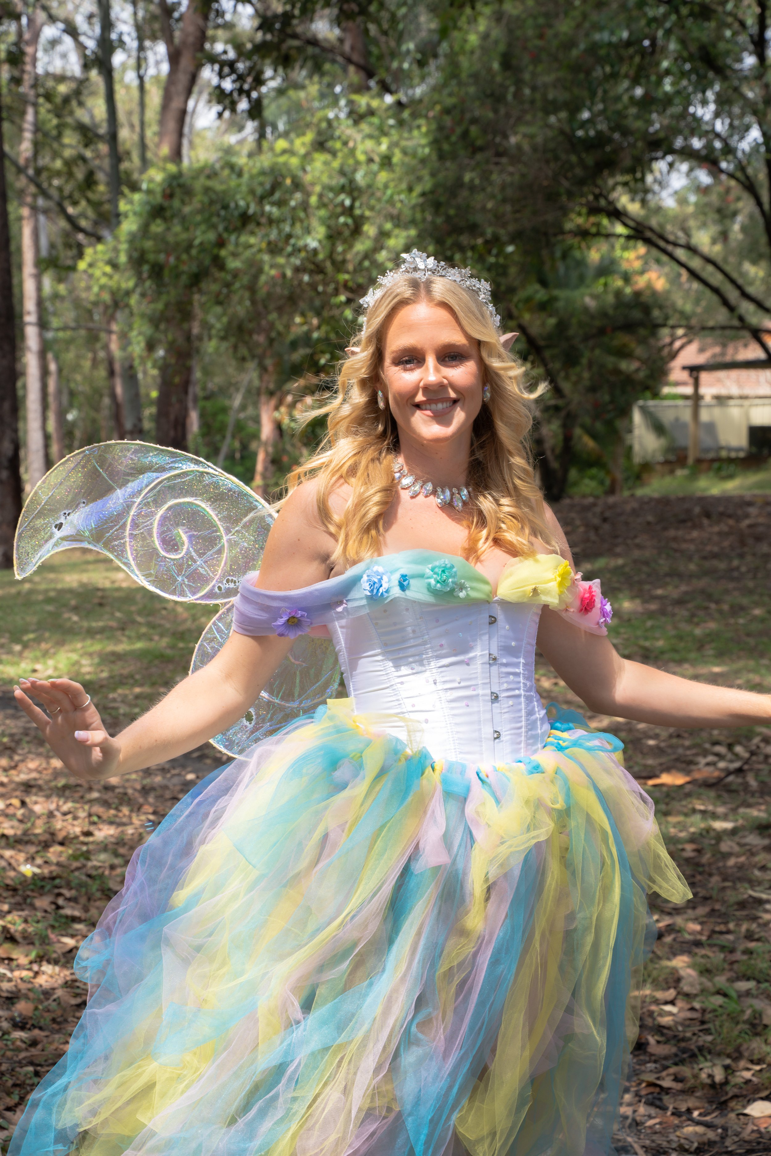A woman dressed as a fairy with blonde hair, wearing a colorful pastel tutu, fairy wings, and a tiara, smiling outdoors in a wooded area.